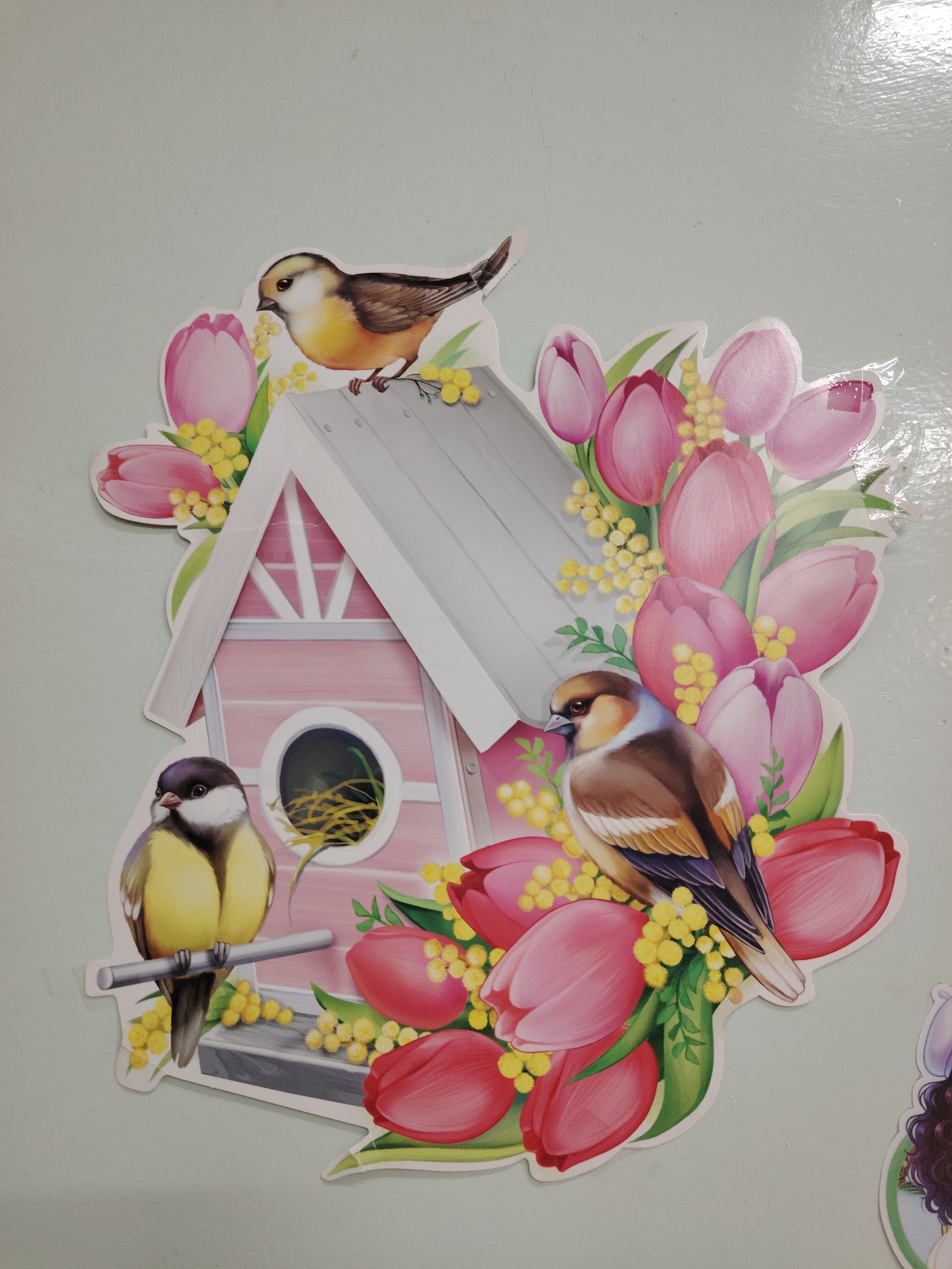 The image depicts a sticker featuring a birdhouse and three birds, surrounded by tulip and mimosa flowers. The birdhouse is pink and white, with a round opening and a roof. One bird sits on the roof of the birdhouse, another is perched on a branch to the left, and the third bird is situated among the flowers. The sticker is placed against a light blue background. There is no information in the image indicating it is in the city of Sarapul, Russia.