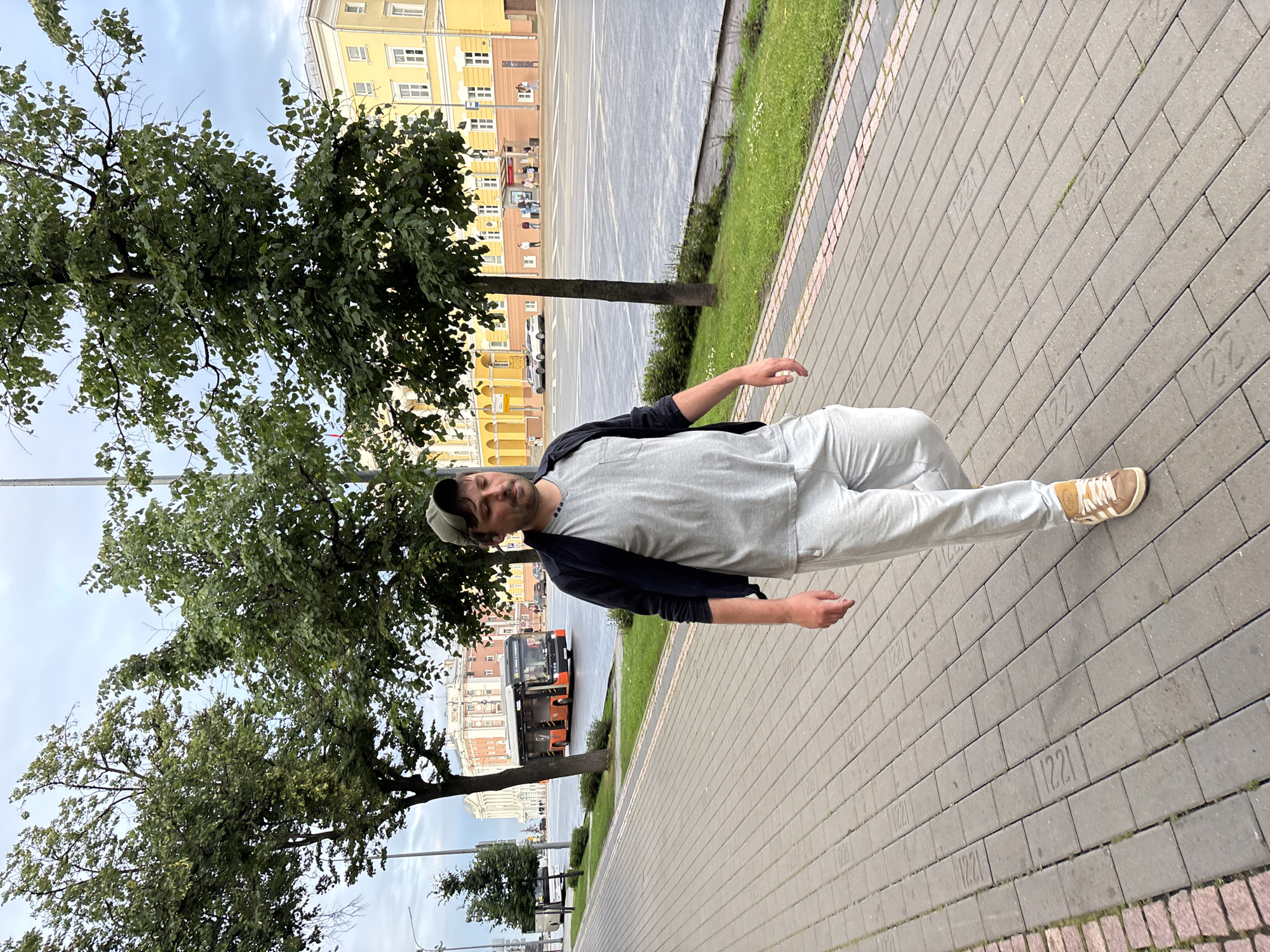 A man walks on a pedestrian sidewalk in Nizhny Novgorod, Russia. He is wearing a dark jacket over a grey t-shirt and light grey sweatpants, with a green baseball cap. He is also wearing brown and white sneakers. The sidewalk is paved with grey rectangular bricks. In the background, there are trees, a street with traffic, including a red and orange bus, and buildings. The sky is overcast.