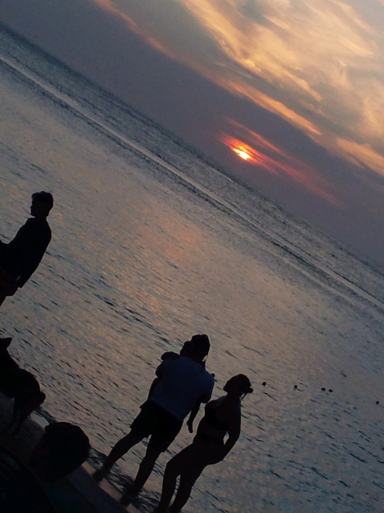 The image depicts a silhouette of a sunset over a body of water. A person stands on the left side, facing away from the camera. In the middle of the frame, a group of people are visible, including one person holding a child. Further to the right, another person is standing. The water appears calm with subtle ripples. In the distance, a few buoys are scattered across the water. The sky is filled with the warm hues of orange and yellow from the setting sun, with scattered clouds creating a dramatic effect. The image is taken from a low angle, emphasizing the vastness of the sky and water.