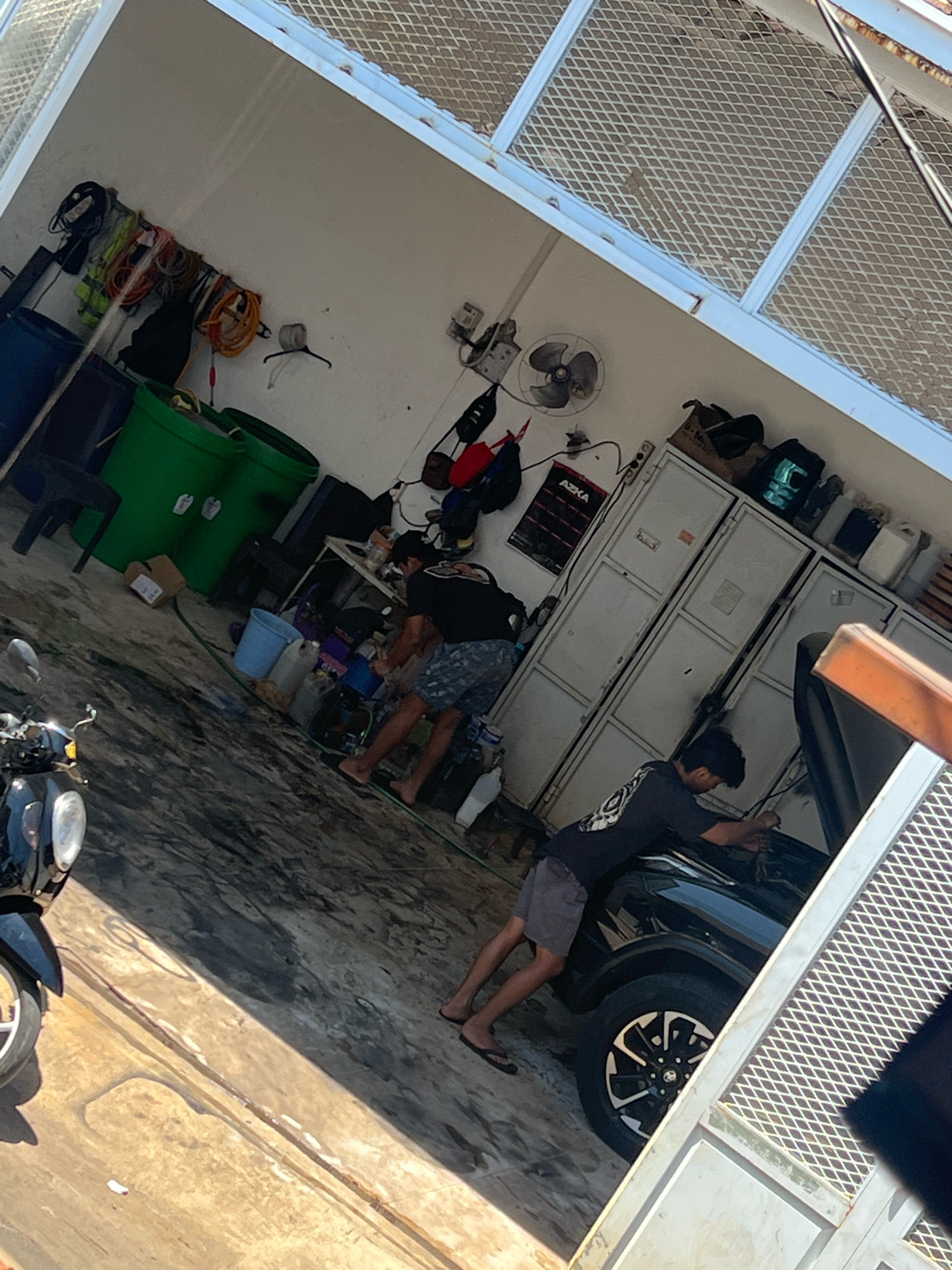 The image depicts an outdoor workshop or garage area, likely in Makassar, Indonesia, characterized by a concrete floor and a partial view of a building with a corrugated metal roof. In the foreground, a black motorcycle is partially visible on the left. The central focus of the scene involves two individuals working on a black SUV with its hood open. One individual is bent over the engine compartment, appearing to be engaged in repair or maintenance. The second individual is positioned near the front of the vehicle, also seemingly involved in the work. Scattered around the workshop are various items, including large green bins, buckets, boxes, tools, and electrical equipment mounted on the wall. The environment suggests a functional workspace, possibly a car repair shop or a private maintenance area.