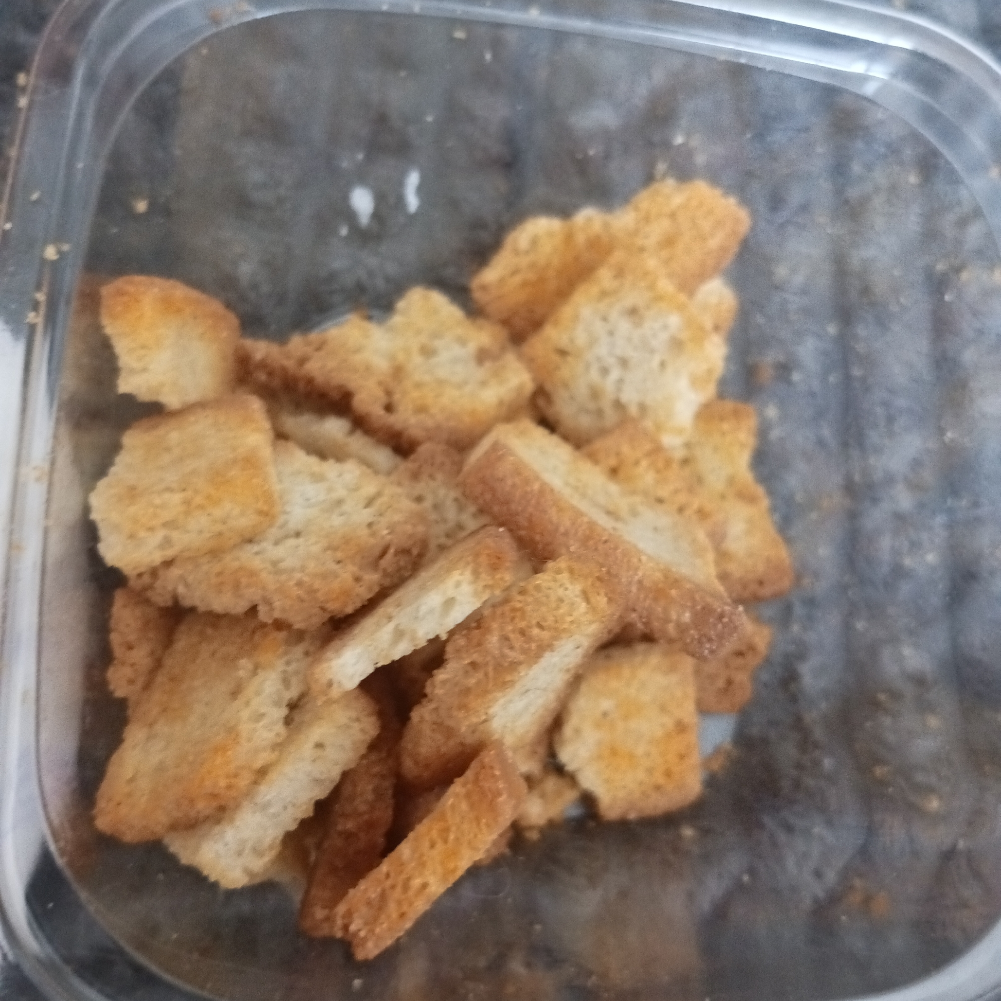 The image presents a close-up, overhead view of a transparent, rectangular plastic food container. Inside, a quantity of golden-brown bread croutons is visible, filling approximately two-thirds of the container's volume. The croutons are irregularly shaped, consisting mostly of small rectangular pieces and cubes, exhibiting a toasted texture and visible internal bread structure. Scattered orange-red seasoning specks are present on their surfaces. The clear plastic container shows minor surface reflections and slight smudges. The background beyond the container is out of focus, appearing as a dark, indistinct, textured surface. The scene is located in the city of Chaikovsky, Russia.