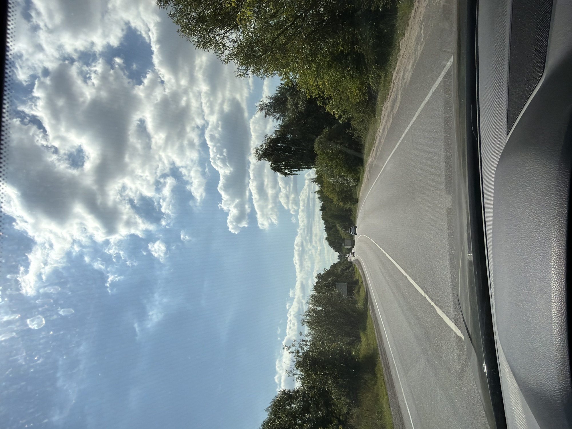 The image depicts a view from inside a vehicle, looking out through the windshield at a two-lane asphalt road curving to the right. The road is marked with white lines and has a grassy shoulder on both sides. Dense green trees line the road, with a few scattered clouds in a blue sky. In the distance, several vehicles, including a truck, are visible on the road. The foreground shows the interior dashboard of the vehicle. The scene appears to be set in a rural or suburban area. The location in Kolontaevo, Russia, is mentioned, suggesting this road is in that region.