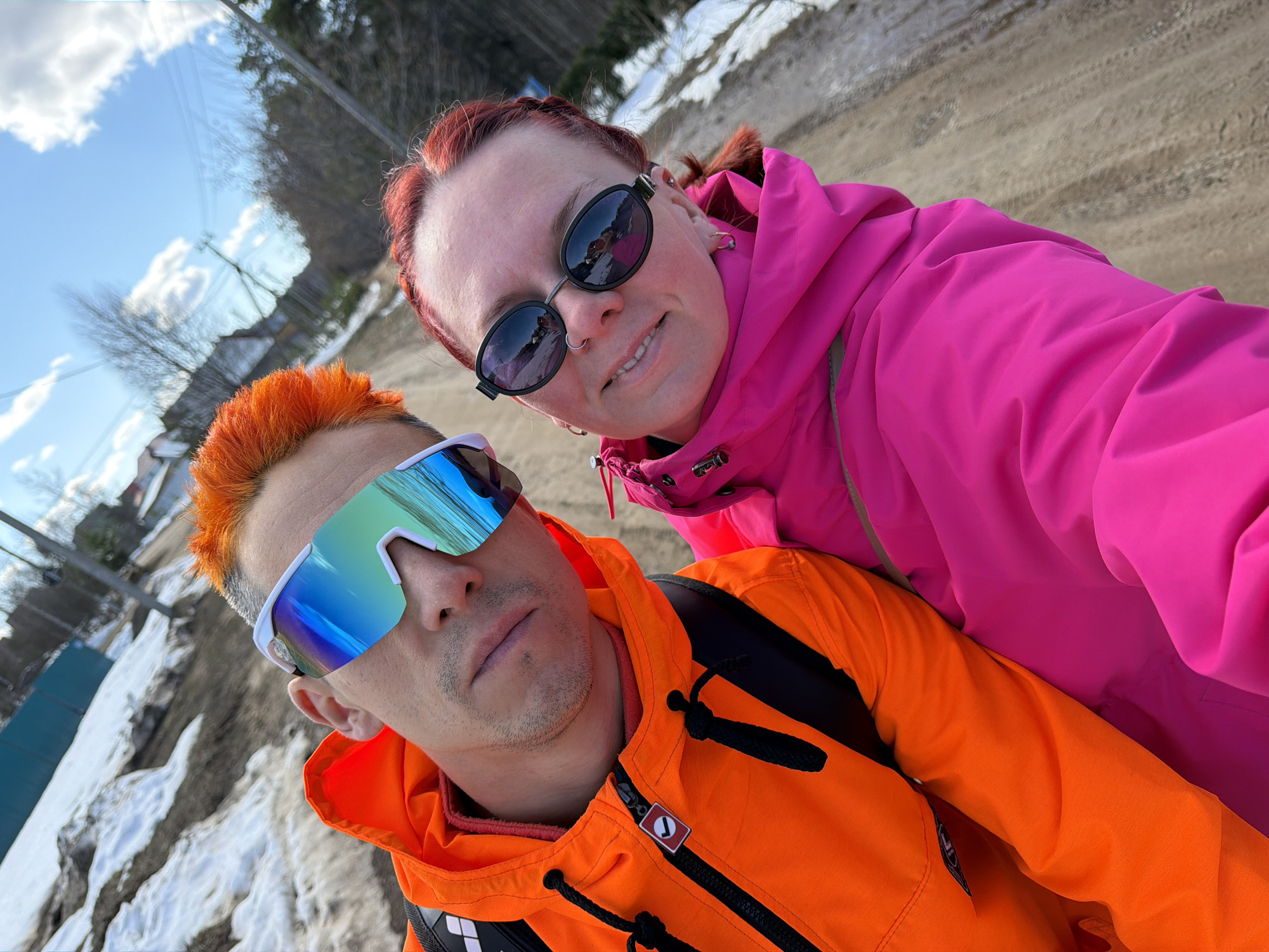 A selfie depicts a man and a woman standing outdoors on a bright day. The man in the foreground has orange hair and wears white-framed sunglasses with reflective blue lenses. He is wearing an orange jacket with black details and a logo visible on the chest. Behind him, the woman wears black sunglasses and a bright pink jacket.
The background suggests a location with some snow on the ground and trees in the distance. It is in the city of Chaikovsky, Russia.