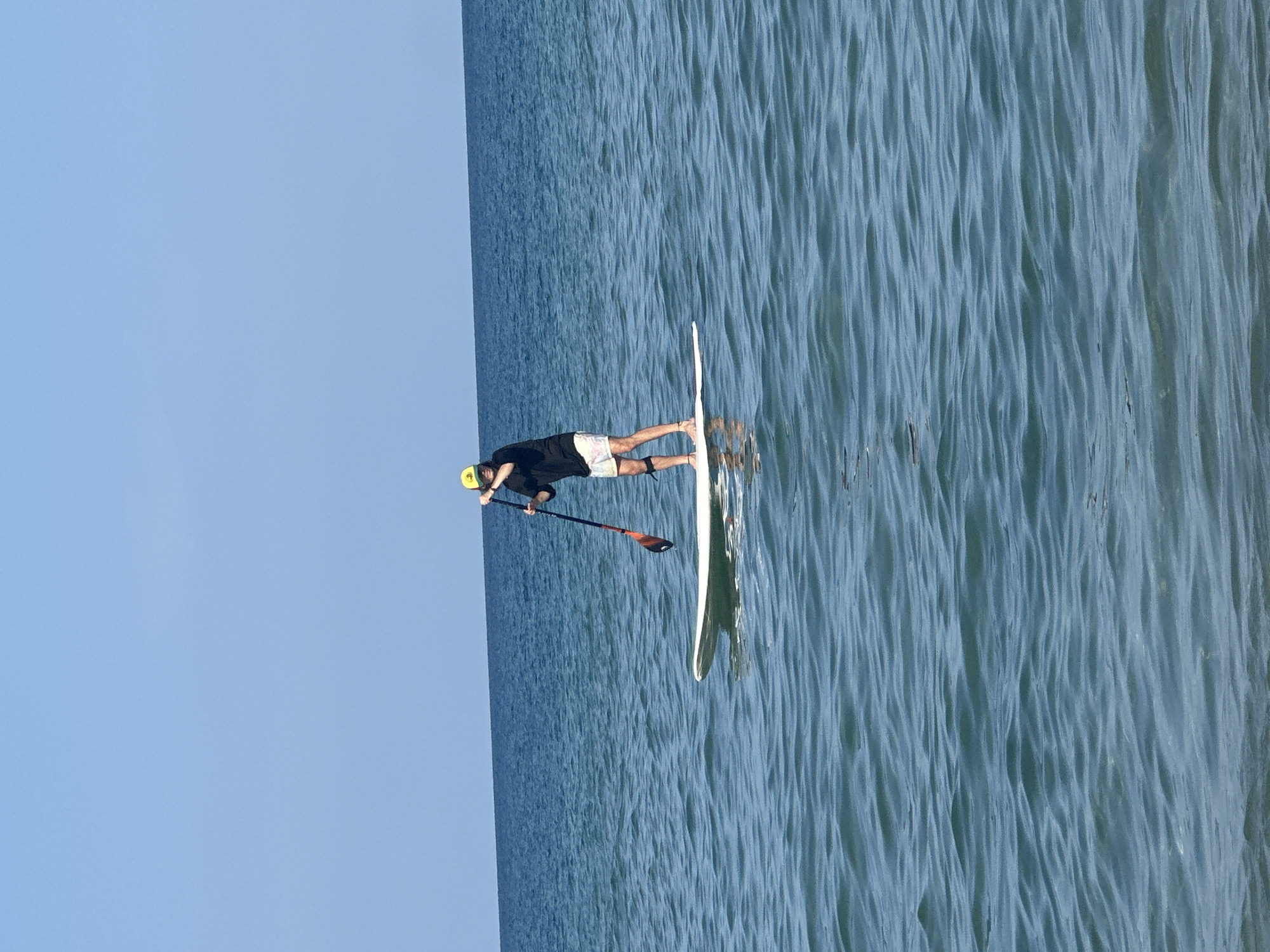 A person stands on a white paddleboard in calm, blue water, holding a paddle. The person is wearing a black long-sleeved shirt, patterned shorts, and a yellow baseball cap. The horizon is visible in the distance, with a clear blue sky above. The scene appears to be a serene day at the beach or lake.