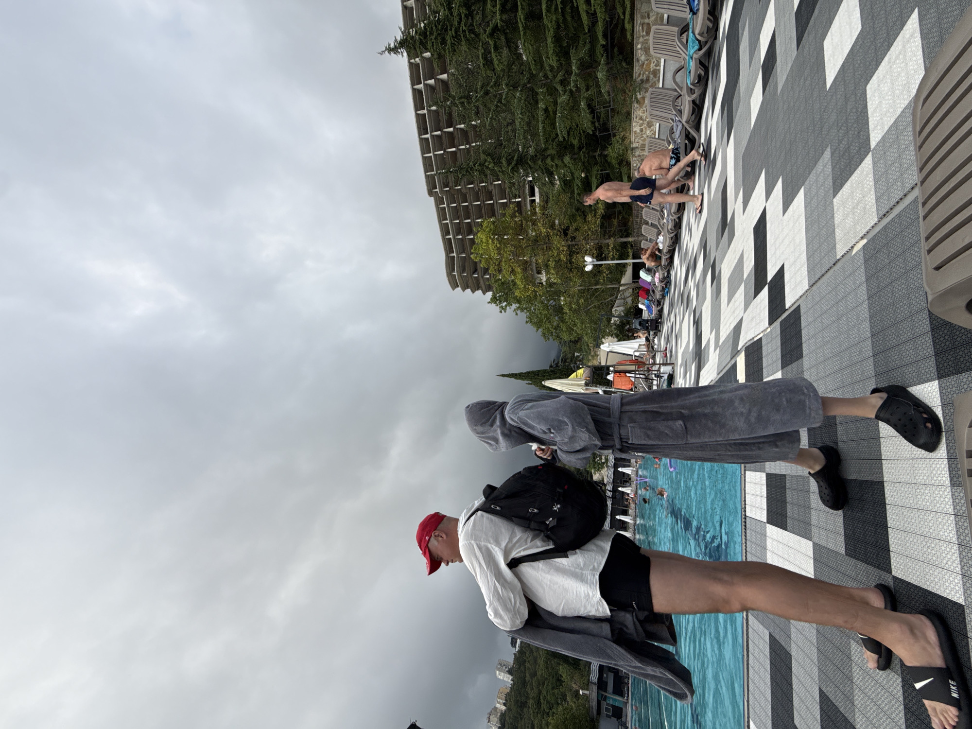 The image depicts an outdoor swimming pool area with a tiled deck. In the foreground, two individuals are standing, one wearing a red cap, white shirt, black shorts, and a backpack, holding a grey towel. The other person is wearing a hooded grey robe. To the right, several people are relaxing on lounge chairs by the pool. In the background, a large, unfinished building stands amidst lush green trees and a hillside. The sky is overcast, suggesting a cloudy day. The scene appears to be in Yalta, Ukraine, possibly at a resort or hotel complex.