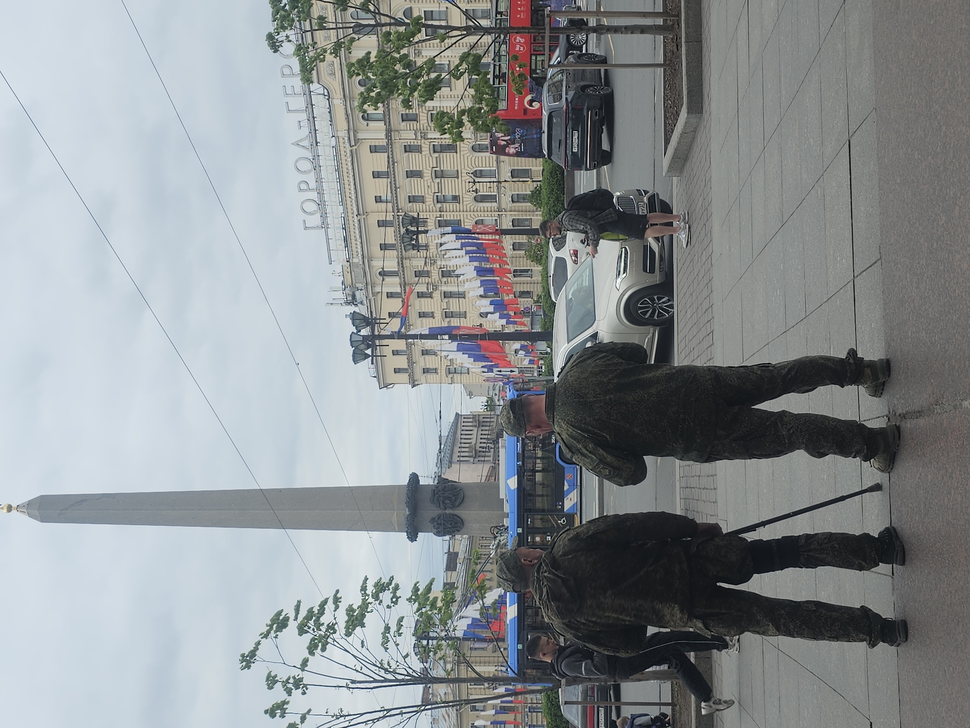 Here is a description of the image:
The image shows a street scene in Saint Petersburg, Russia. In the background is the Alexander Column in Palace Square. The Winter Palace is not visible, but the General Staff Building is. Decorations of Russian tricolor flags are visible. Two men in military fatigues are visible on the sidewalk, possibly engaged in crowd control. A white SUV is parked on the street. The sign "GOROD-GEROY" (Hero City) on the building behind the SUV.