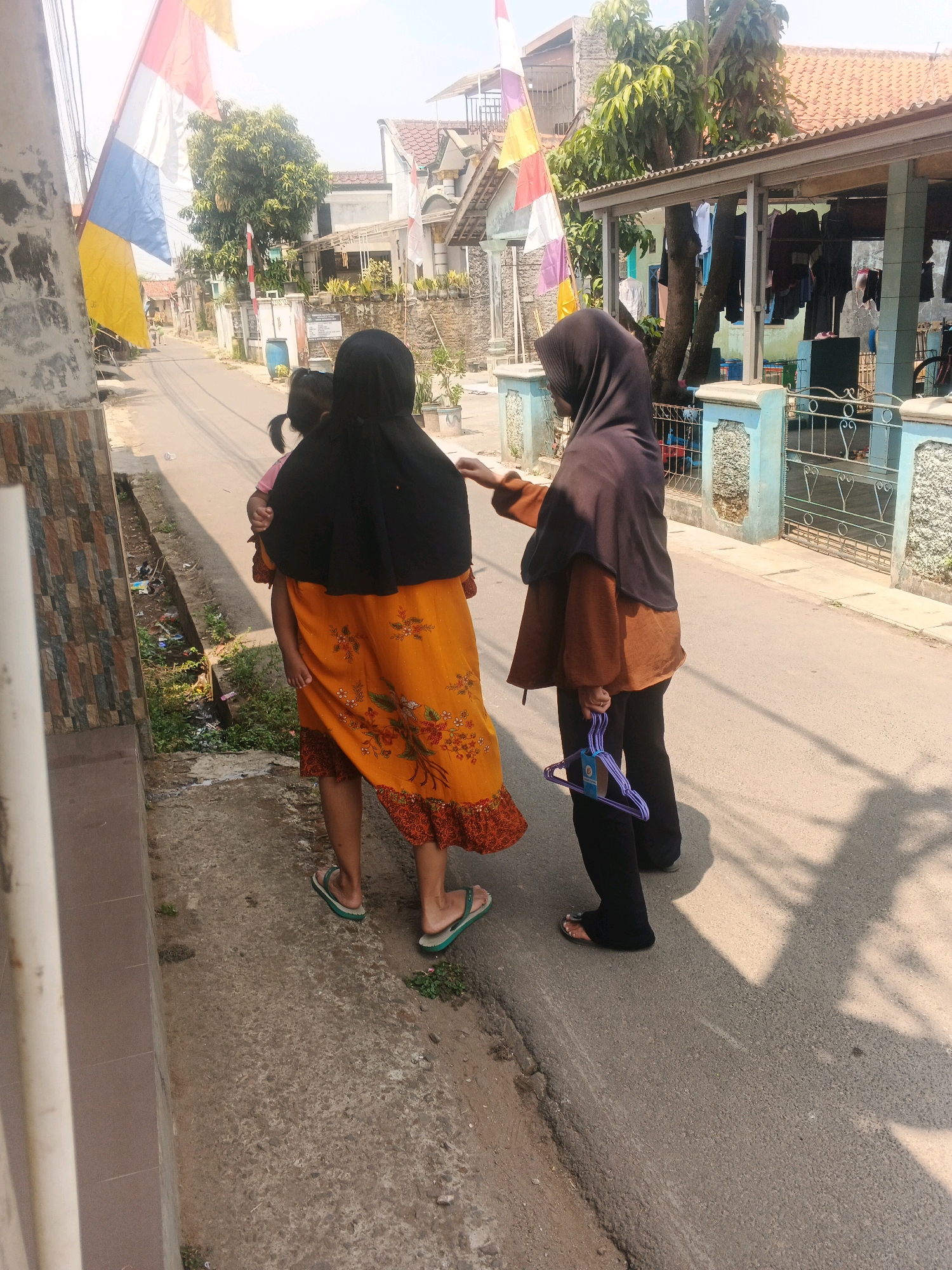 In Kadu, Indonesia, two women and a child are walking on a paved street. The woman on the left is wearing a black hijab and an orange dress with a floral pattern. She is holding a child who is facing away from the camera. The woman on the right is wearing a black hijab and a brown top with black pants, and she is carrying a purple clothes hanger. The street is lined with houses and trees, and there is a fence on the right side of the image. The sun is shining brightly, casting shadows on the ground. There are flags hanging on poles on the left side of the image. The scene depicts a typical day in a residential area.