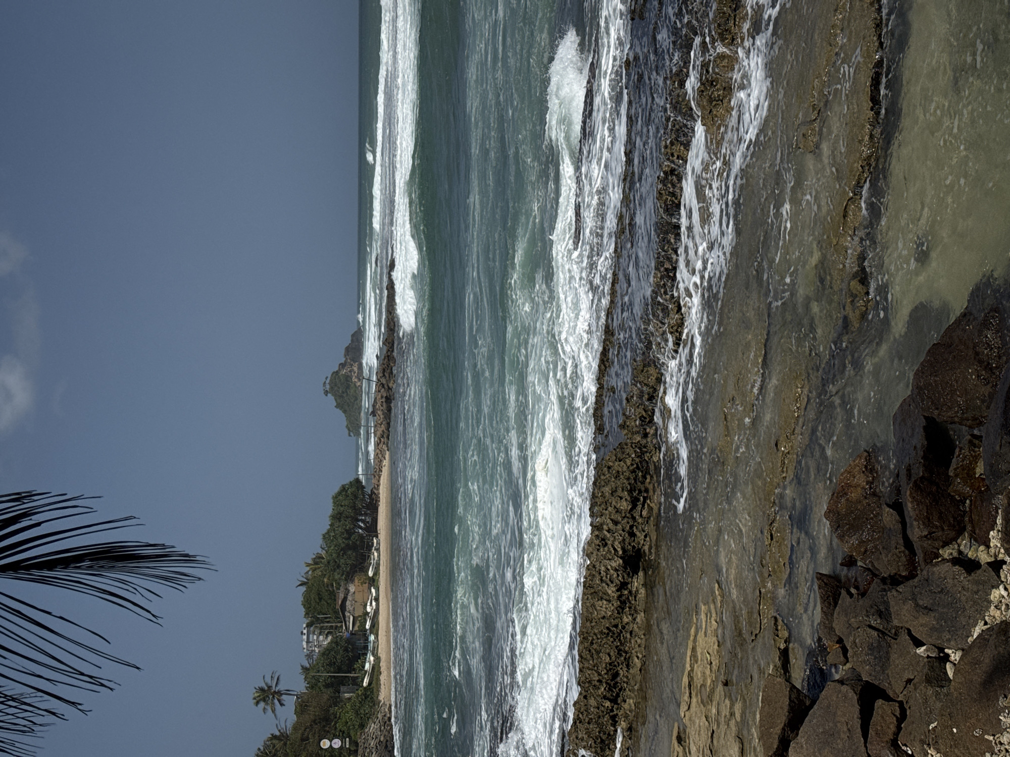 In Ahangama, Sri Lanka, a rocky coastline meets the turquoise waters of the ocean. The foreground is dominated by large, dark brown and grey rocks, with smaller pebbles scattered amongst them. These rocks form a natural barrier, with clear, shallow water pooling in the crevices and flowing over the flatter surfaces.
Beyond the rocky shore, the ocean is a vibrant blue-green, with white-capped waves rolling towards the land. The surf is moderate, creating foamy patterns as it breaks against further submerged rocks. In the distance, a small, rocky island with a lone tree stands against the horizon, adding a focal point to the seascape.
The middle ground reveals a sandy beach dotted with several small, white boats pulled ashore. Behind the beach, a dense line of lush green trees and palm trees forms a natural backdrop. A few buildings, appearing to be accommodations, are visible nestled amongst the foliage. A single, dark palm frond hangs into the upper left corner of the frame, its shadow partially obscuring the clear, pale blue sky. The overall scene depicts a serene coastal landscape on a bright, sunny day.