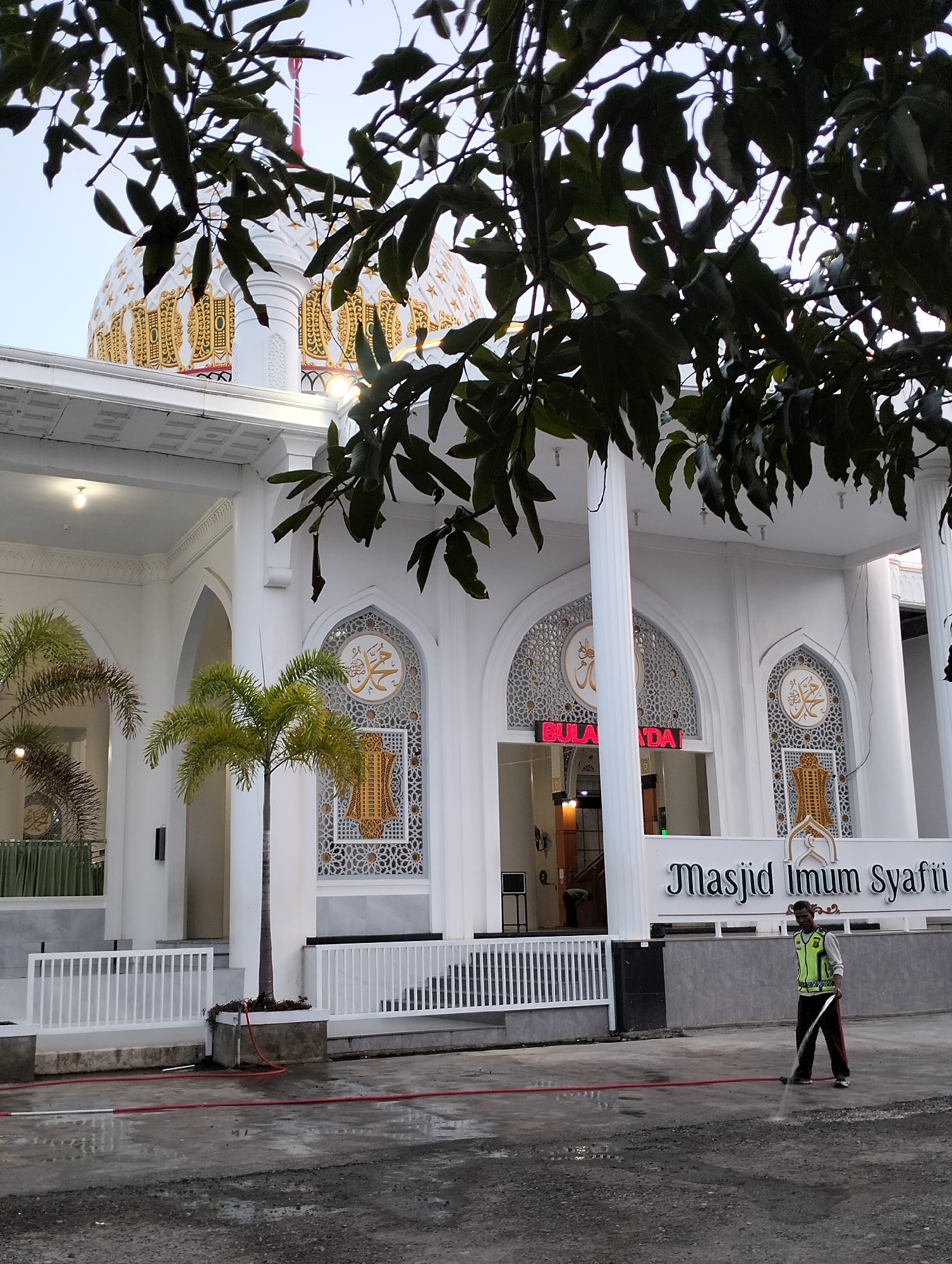 An exterior shot of a mosque with a white facade and a gold-domed roof is captured in Blangmetimu, Indonesia. The mosque features intricate gold embellishments on its dome and arched windows, which contain Arabic calligraphy. In front of the mosque, a man wearing a bright green safety vest and dark pants is hosing down the wet pavement with a red hose, appearing to be cleaning the area. Several palm trees are present on the mosque grounds, and lush green foliage from overhanging trees partially obscures the view of the upper portion of the building. The sky is overcast, suggesting it might be early morning or late afternoon. A white railing separates the paved area from the mosque's entrance. The name of the mosque, "Masjid Imam Syafii," is visible on a white sign.
