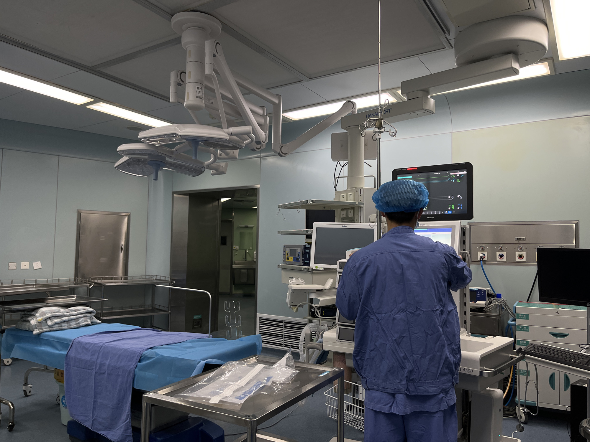 The image depicts the interior of a medical facility, likely an operating room or an intensive care unit, characterized by its sterile and equipped environment. The room features light blue walls and a white ceiling with recessed rectangular lighting panels.
In the foreground, a medical professional, seen from the back, is wearing blue scrubs and a blue surgical cap. They are standing in front of a complex array of medical equipment, including several monitors displaying vital signs and other medical data. One monitor clearly shows the brand "PHILIPS". The individual appears to be interacting with the equipment, possibly adjusting settings or reviewing information.
The room is equipped with various specialized medical devices. Overhead, multiple surgical lights with articulated arms are suspended from the ceiling, along with a robust ceiling-mounted system labeled "HANAUFORT", which supports additional monitors and an IV pole. To the left, a surgical bed is prepared with blue disposable covers and folded blankets, suggesting readiness for a patient or post-procedure recovery. Nearby are stainless steel trolleys, one holding clear plastic-wrapped medical supplies.
On the right side of the image, an anesthesia machine, possibly a "Dräger A500" (partially visible), is connected to the ceiling-mounted system. There are also wall-mounted outlets for medical gases or power, and another workstation with a black monitor and keyboard, featuring cabinets labeled with Chinese characters such as "第一层" (first layer) and "第二层" (second layer). A metal door and additional stainless steel shelving units are visible along the left wall. The overall impression is one of a functional, well-equipped, and clinically clean medical space. No specific time of day or emotions are discernible. The sterile environment and specialized equipment serve as strong location cues for a medical setting.