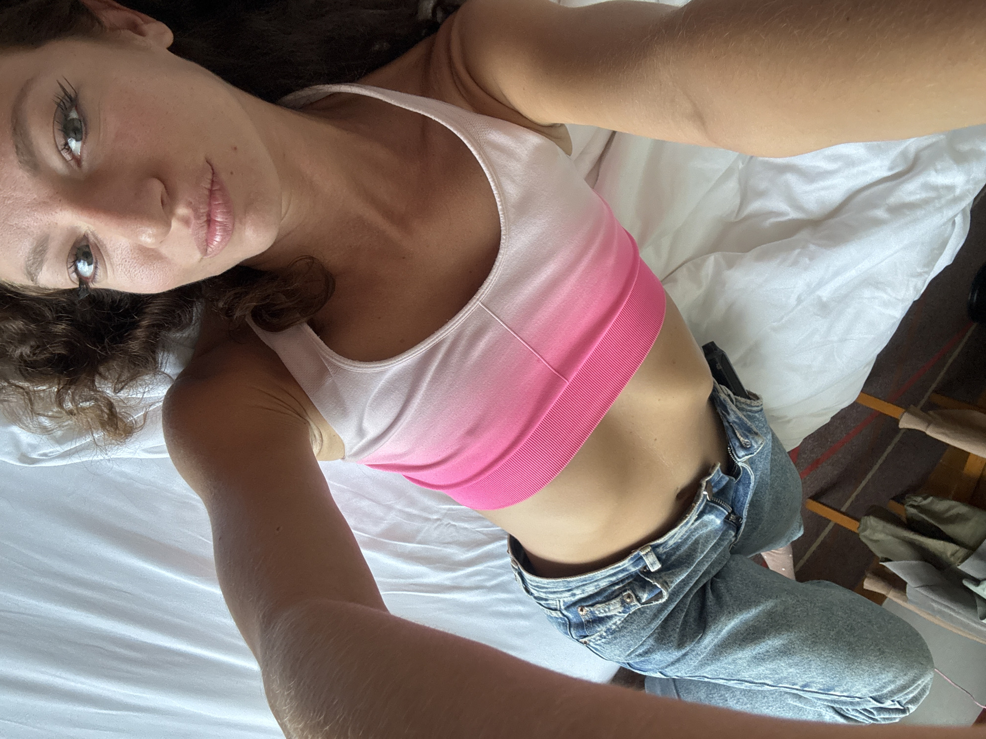 A woman is lying on a white bed, wearing a pink and white ombre crop top and blue jeans. She is looking at the camera. The location is unspecified; there is no indication it is Đà Nẵng, Vietnam. A closet is partially visible in the background with some items of clothing.