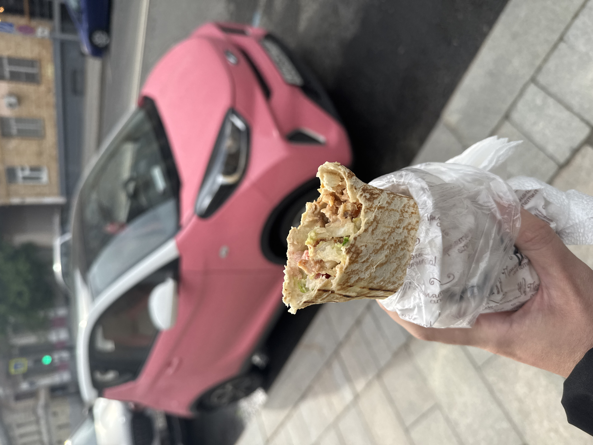 A person holds a partly eaten shawarma wrapped in plastic, standing outside in Moscow, Russia. A pink car with white accents is visible in the background. The ground is paved with rectangular stones.