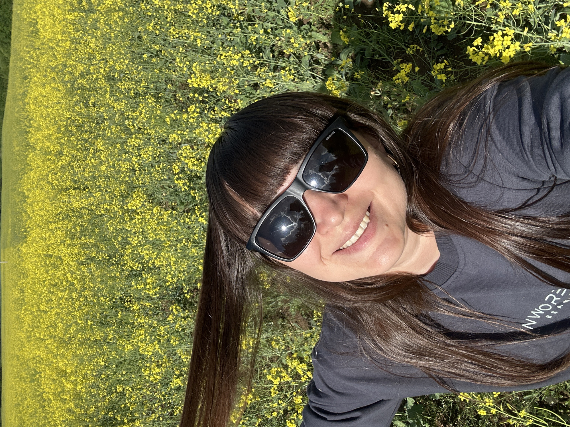 A woman stands in a field of yellow flowers, likely rapeseed or canola, in an outdoor location. She is wearing sunglasses and a gray long-sleeved shirt with a white logo or lettering visible. Her hair is dark brown and straight, with bangs. She is smiling. The photo appears to be taken from a low angle, looking up at her. It is difficult to precisely pinpoint the location within Sarapul, Russia, based on the given image, as the surroundings only show the field of flowers.