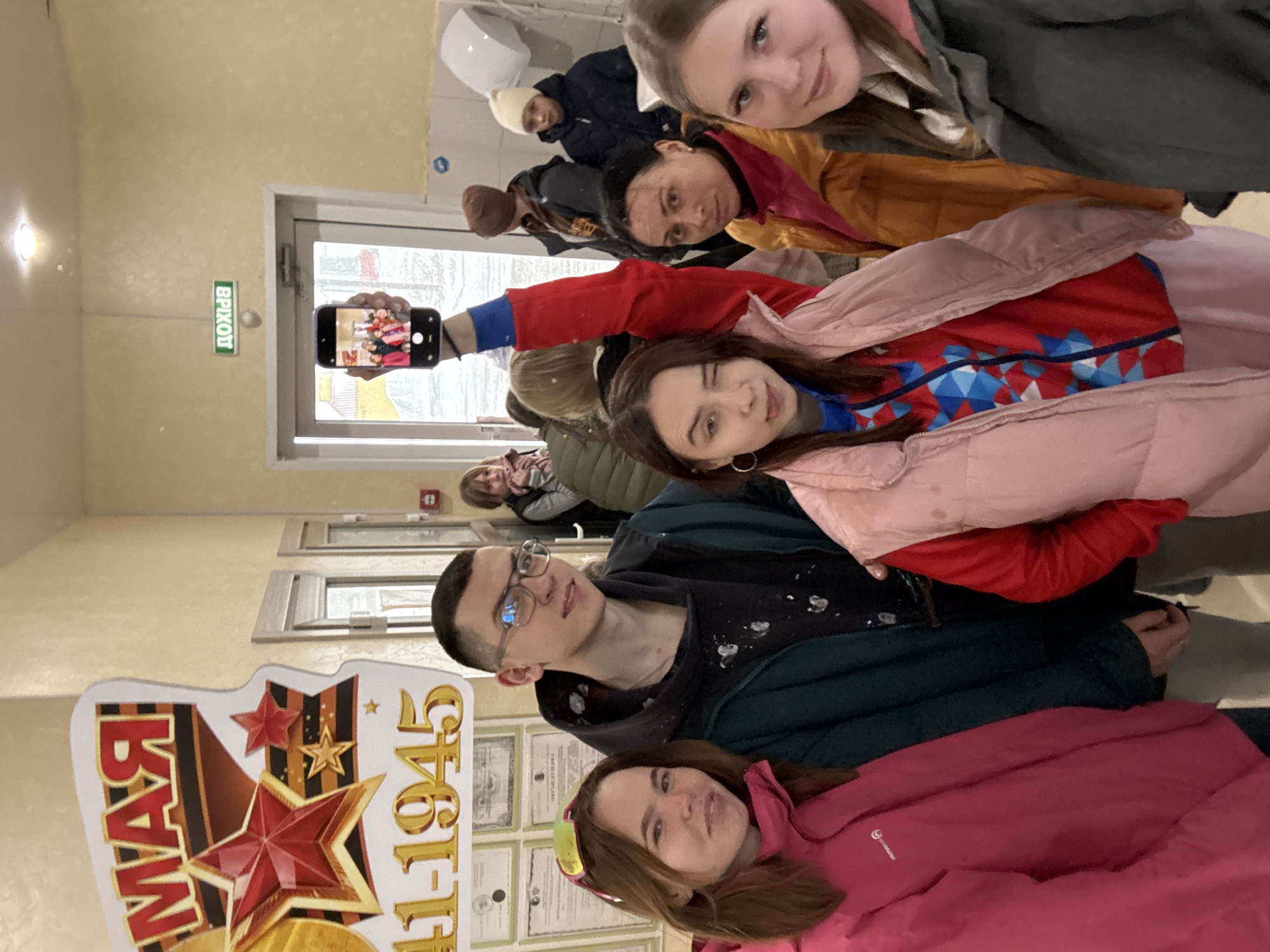 A group of people is taking a selfie in a hallway, with an additional group of people visible in the background standing near a doorway. The people in the foreground are posing, with one individual holding a phone up to capture the group. There is a sign with red stars and text that includes "1945" on the left side of the frame. Certificates or documents are visible in the background. The location is potentially Gory, Russia. The people are wearing jackets.