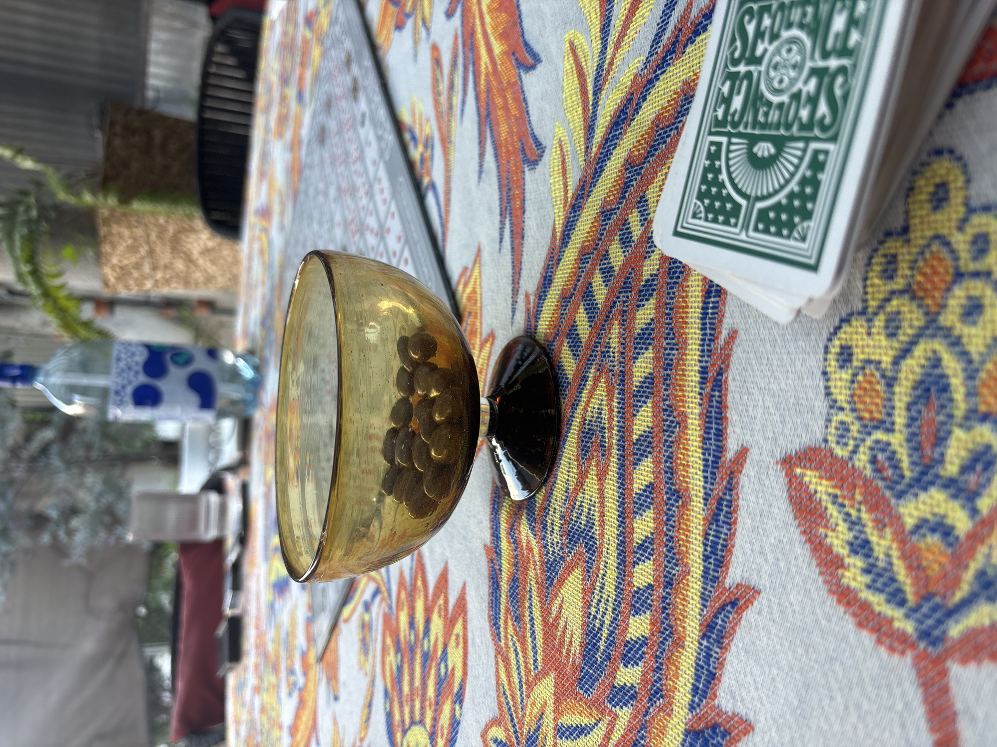 The scene depicts a table setting with various objects on a patterned tablecloth. An amber-colored glass with a dark base contains what appear to be small round objects. A stack of playing cards with the word "Sequence" printed on the top card is also present. In the background, there is a water bottle, a blurred view of foliage, and structures suggesting an outdoor setting. The location is Puebla, Mexico.