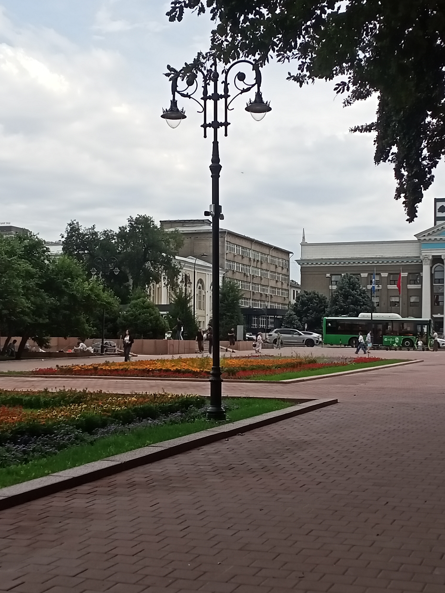 The image shows an outdoor urban scene in Bishkek City, Kyrgyzstan, under an overcast sky. In the foreground, a wide paved area with a brick pattern leads to a landscaped area with flowerbeds containing orange, red, yellow, and green foliage. A dark lamppost with intricate metalwork and two lamps stands prominently in the middle ground, to the right of the flowerbeds. Behind the flowerbeds, several people are visible in a park-like setting with trees. Further in the background, a green bus is driving on a road, and beyond that, several buildings are visible, including a multi-story building with many windows on the left and a larger building with columns and flags on the right. One of the flags appears to be the flag of Kyrgyzstan. The scene depicts a public space with elements of urban infrastructure, landscaping, and pedestrian activity.
