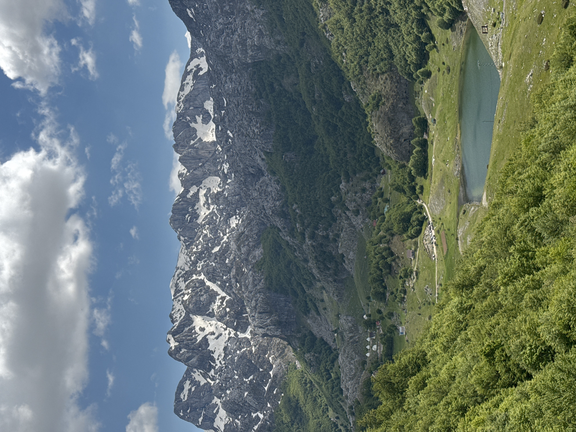 An eye-level view of a mountain landscape in Brskut, Montenegro. A tall, rocky mountain with patches of snow stands adjacent to a green, forested slope that leads down to a small lake. A cluster of buildings, likely residences or tourist accommodations, are visible near the base of the slope. Above the mountain is a blue sky with scattered white clouds.