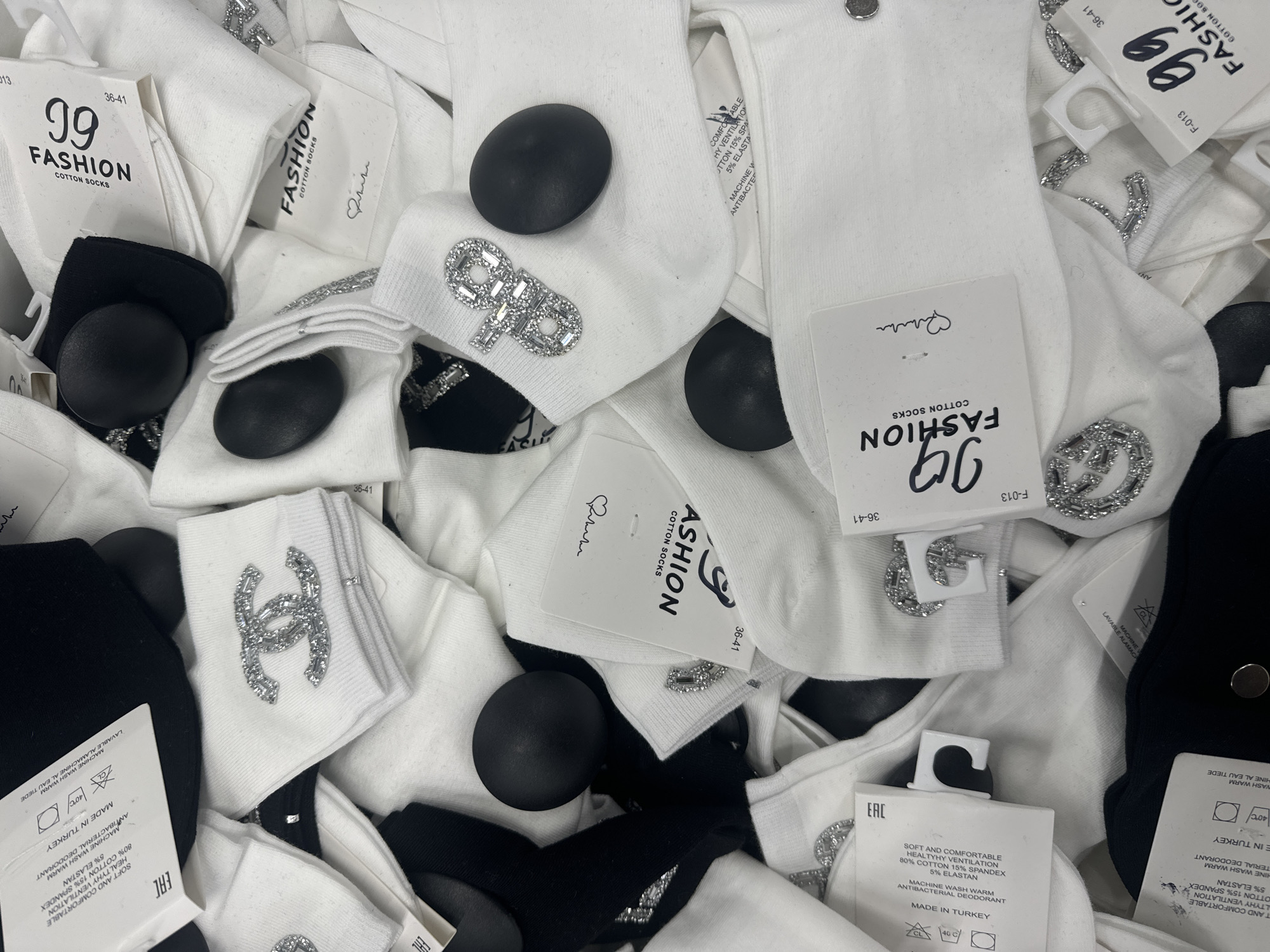 The image shows a pile of white cotton socks with various brand logos and embellishments, including a Chanel-like interlocking "C" design and a logo resembling "GP" spelled out in crystals. Some socks are partially black. Tags indicate the socks are made of 80% cotton, 15% spandex, and 5% elastane, and were made in Turkey. Black round objects, possibly decorative studs, are interspersed among the socks.