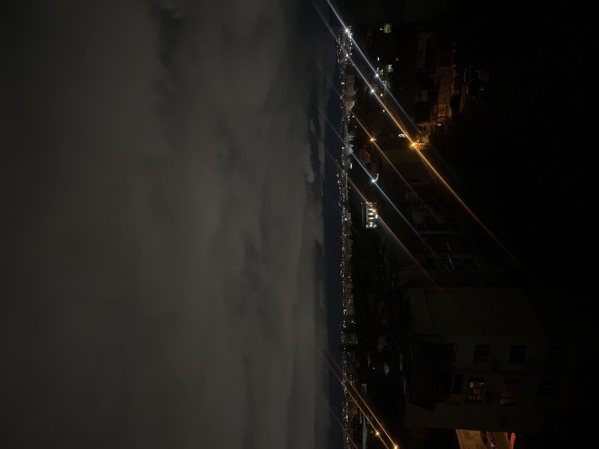 This media file presents a nighttime view of Sofia, Bulgaria, captured from a high vantage point, looking out over the city. The image is oriented sideways, but when rotated correctly, the sky occupies the upper portion, and the city lies below.
The top half of the frame is dominated by a dark, cloudy sky. The clouds appear dense and textured, obscuring most celestial bodies, though faint specks of light are visible in some clearer patches.
Below the cloudy expanse, the city stretches across the horizon, marked by a dense array of artificial lights. These lights, emanating from buildings and street infrastructure, form a bright, continuous line across the middle of the image. Many of these light sources produce distinct, radiating streaks of light, appearing in white, blue, orange, and yellow, creating a starburst effect.
In the foreground, towards the bottom of the image, the dark silhouettes of buildings and structures are visible. Some of these structures have illuminated windows, offering glimpses of interior lights. The details of these foreground buildings are largely obscured by the darkness, but their general shapes and outlines are discernible.
The overall scene depicts a serene yet illuminated cityscape under a cloudy night sky. There is no human activity or visible text in the image.