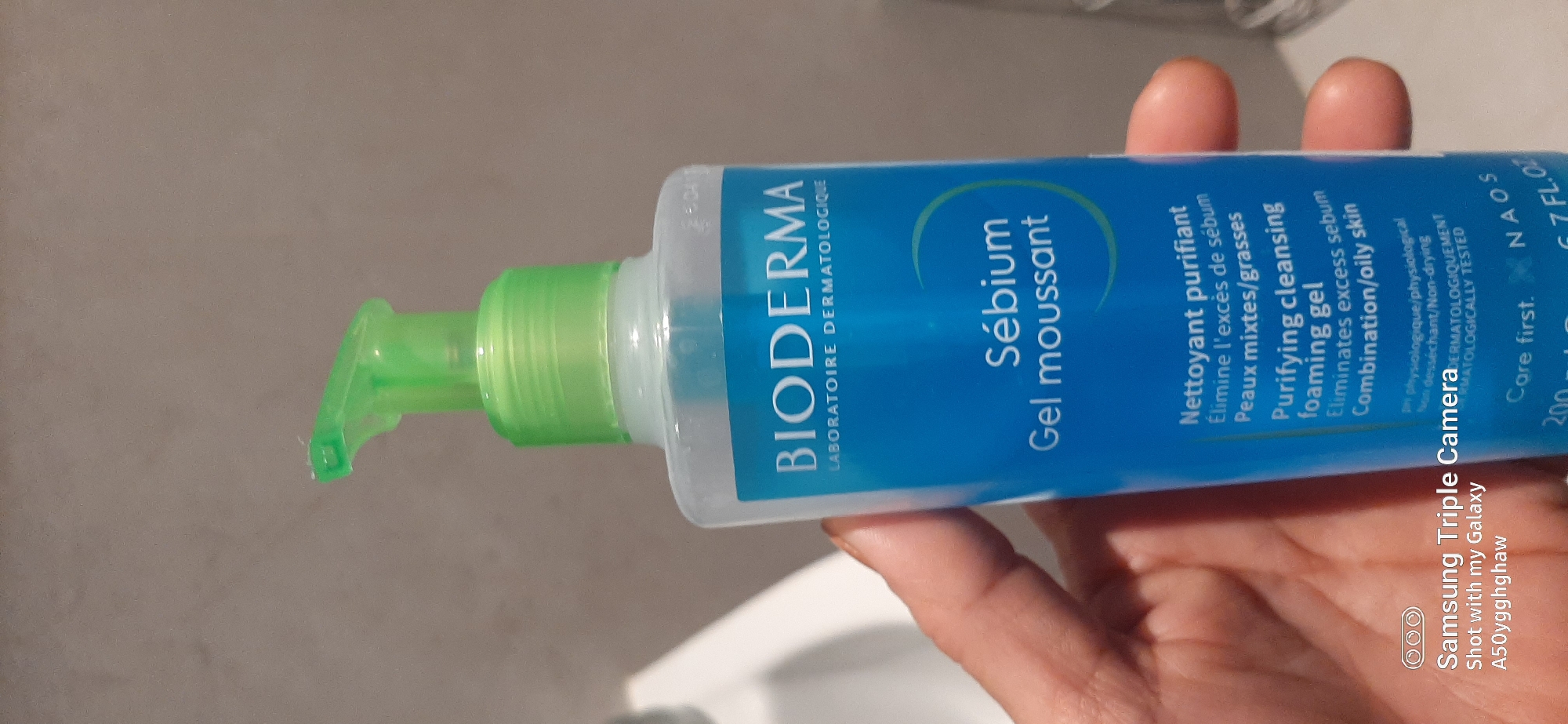 The image shows a close-up of a person's hand holding a bottle of Bioderma Sebium Foaming Gel. The bottle is clear with a blue label and a bright green pump dispenser. The label has text in French and English, indicating it is a "Purifying cleansing foaming gel" for "Combination/oily skin." The background is out of focus and appears to be a tiled wall, possibly in a bathroom. The bottom of the image contains a watermark that reads "Samsung Triple Camera Shot with my Galaxy A50yyghghaw." There is no indication that this scene is located in Dubai, United Arab Emirates.