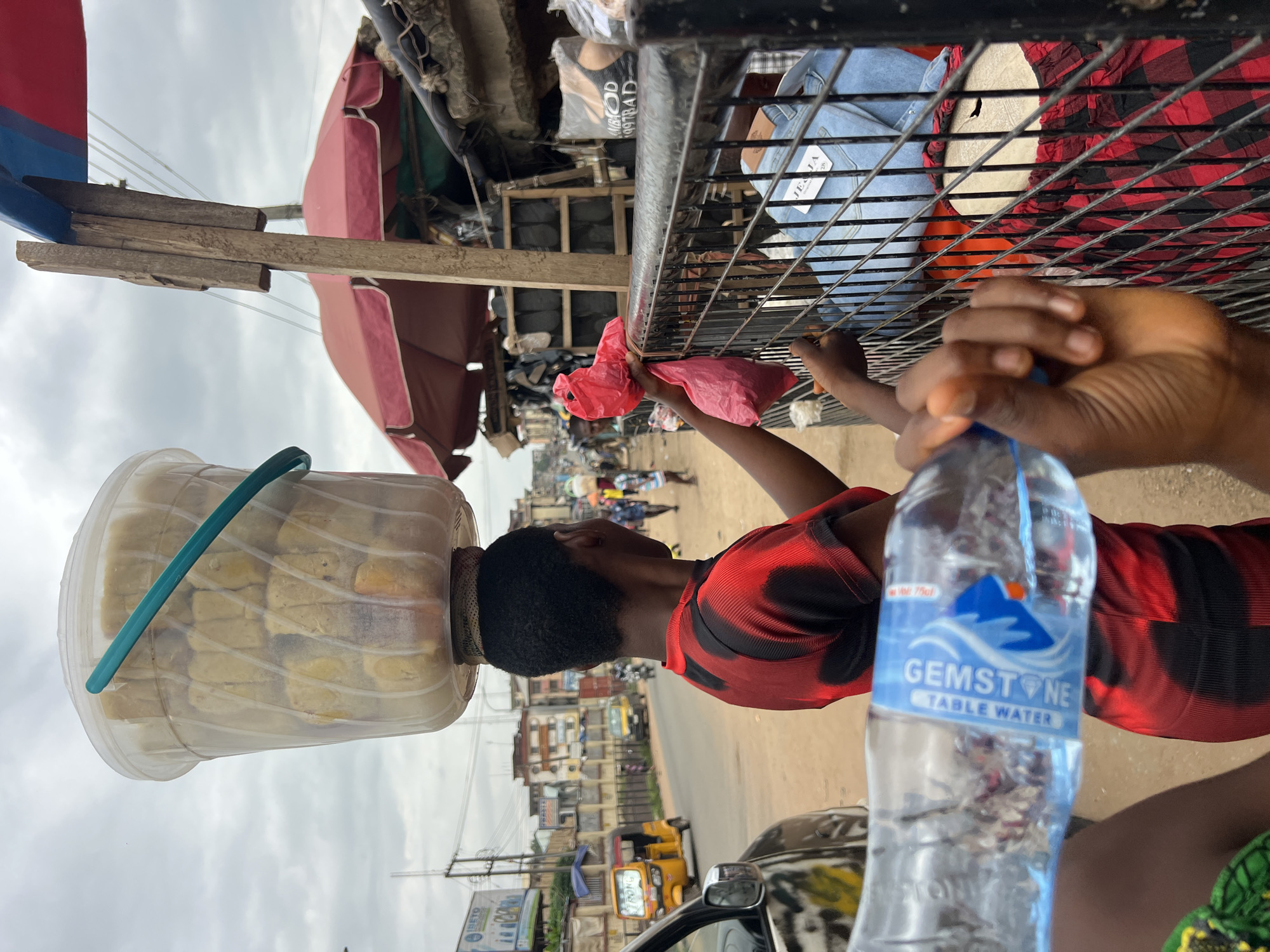 In Nkpor, Nigeria, a vendor balances a large, clear plastic bucket filled with food items on her head, carrying it over a busy street. The bucket is secured by a turquoise strap. She is wearing a red and black patterned shirt. In the foreground, a hand holds a bottle of "Gemstone Table Water." The vendor is interacting with someone behind a metal fence, holding out two pink plastic bags. Behind the fence, a person wearing blue jeans and a red and black checkered skirt is visible, along with various goods displayed at market stalls. The background shows a typical urban street scene with buildings, a yellow auto-rickshaw, and other vehicles and pedestrians. The sky is overcast.