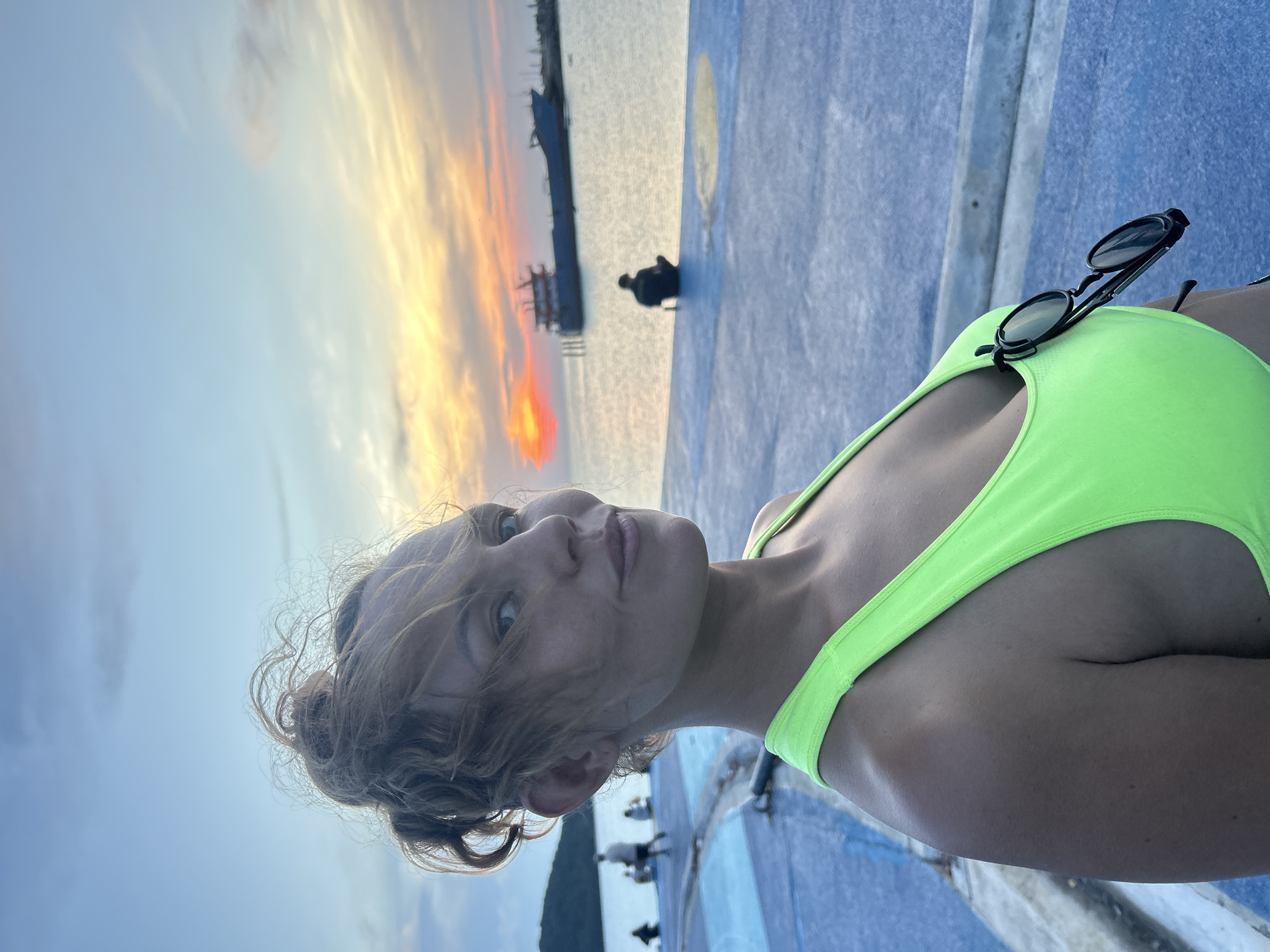 The image shows a woman wearing a green top. Sunglasses rest on the top. The background reveals a sunset over the sea, with silhouettes of structures and possibly people in the distance. The foreground is a blue and white textured surface. The image was taken outdoors, likely at a pier near Thong Sala, Thailand.