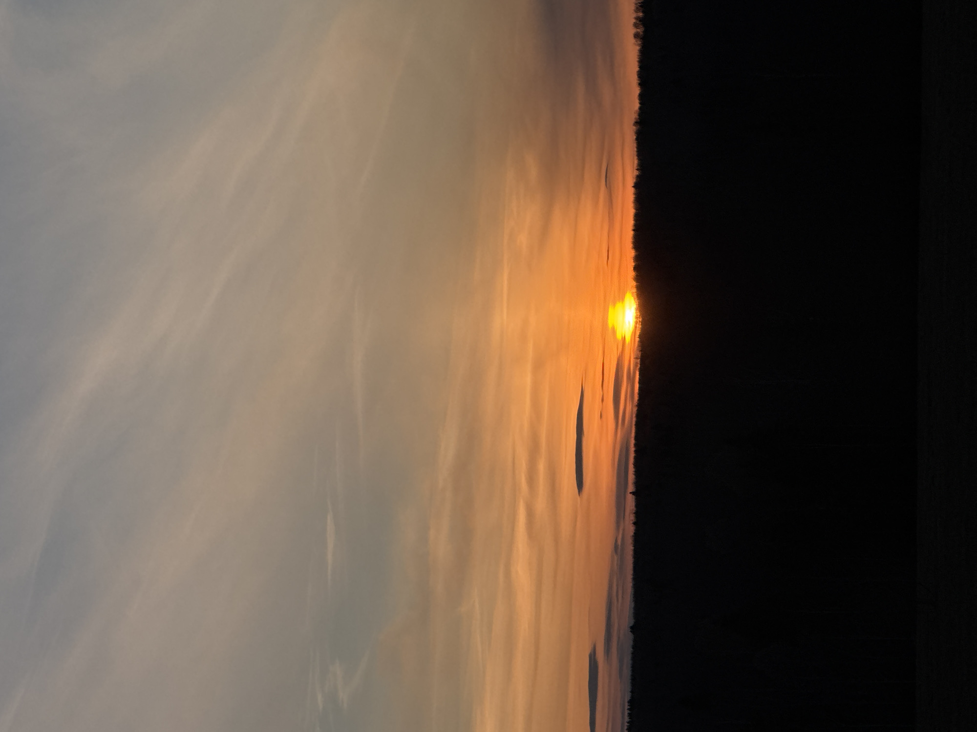 The image shows a sunset view. The sun is positioned near the horizon, emitting a bright orange glow. The sky displays a blend of colors, transitioning from gray-blue towards the upper left to orange closer to the horizon. Dark shapes, possibly trees or land, are silhouetted against the right side of the sky. The image location is not specified, but it has a city name specified, Amin'kai, Russia. There are no people visible in the image. The scene is still and reflects a moment in time as the sun sets.