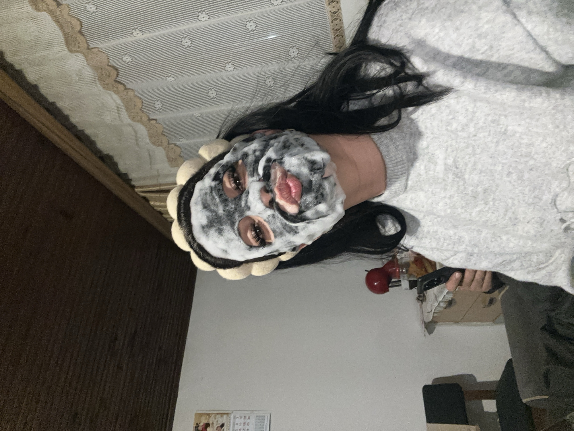 A person with a facial mask is lying in bed. The person has long, dark hair and is wearing a gray sweater. They are holding a black object. The location is a room with white walls and a wooden bed frame. There is also a calendar on the wall. The city of Chubra, Bulgaria is not directly identifiable in the image.