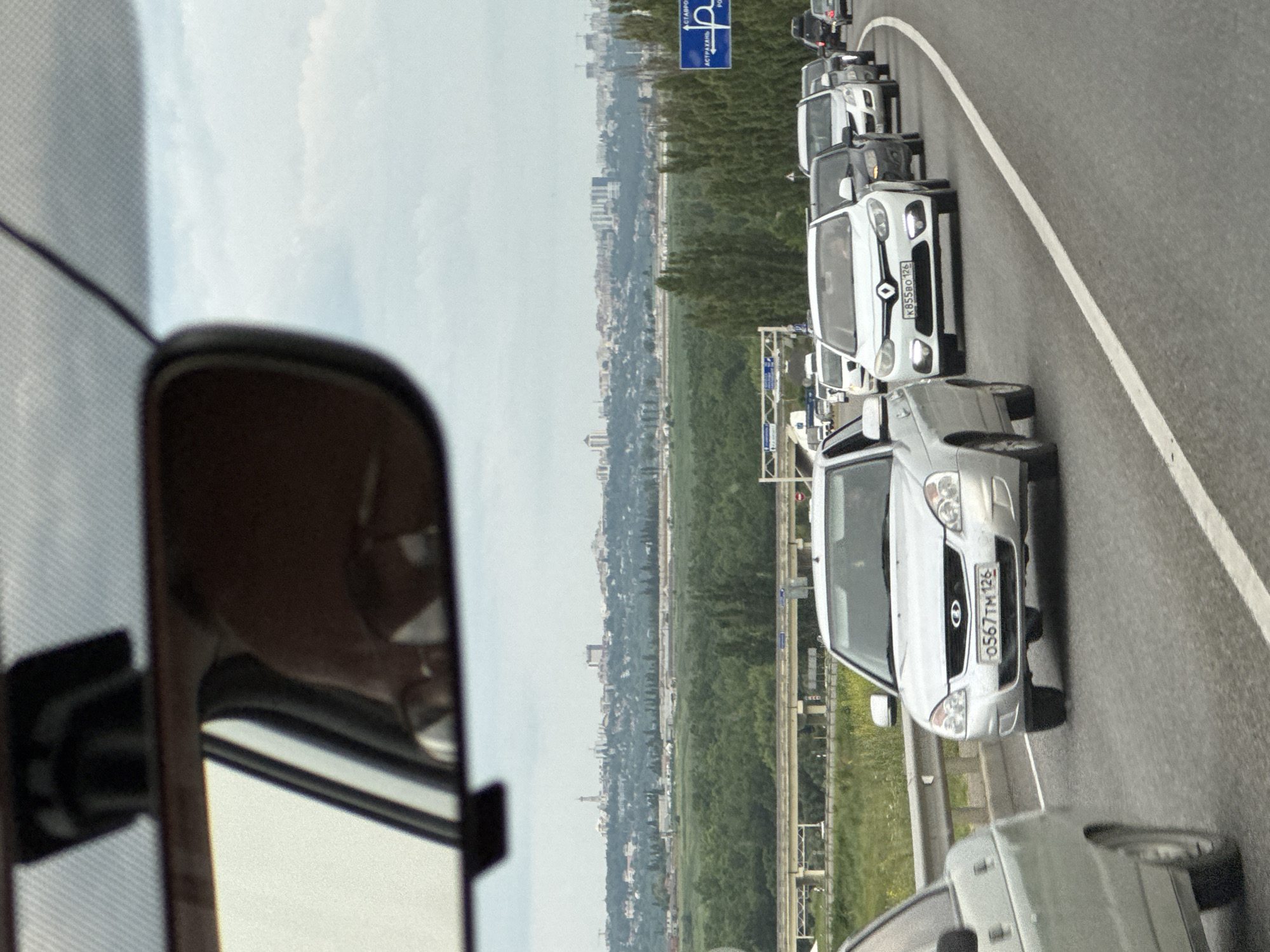 The image shows a traffic jam on a highway near Mikhaylovsk, Russia. A line of cars is visible on the road, with a city skyline and green trees in the background. The sky is overcast, suggesting a cloudy day. The perspective is from inside a vehicle, looking forward and slightly to the side, with a rearview mirror visible in the foreground.
The main subjects are the cars, which appear to be a mix of sedans and vans. Several vehicles have visible license plates. One license plate on a silver car reads "0567TM 126". Another license plate on a white car reads "K 855BO 126".
A blue traffic sign is also visible, with Russian text and an icon of a parking symbol with a wheelchair, possibly indicating accessible parking. The text on the sign is partially visible and reads "АЭРОПОРТ" (Airport) and "РОС" (likely part of a place name).
The overall scene depicts a common occurrence of traffic congestion on a highway, likely during daytime given the ambient light. There are no discernible emotions conveyed as the image focuses on the objects and the scene itself. The presence of Russian license plates and text on the sign confirms the location context as Russia.