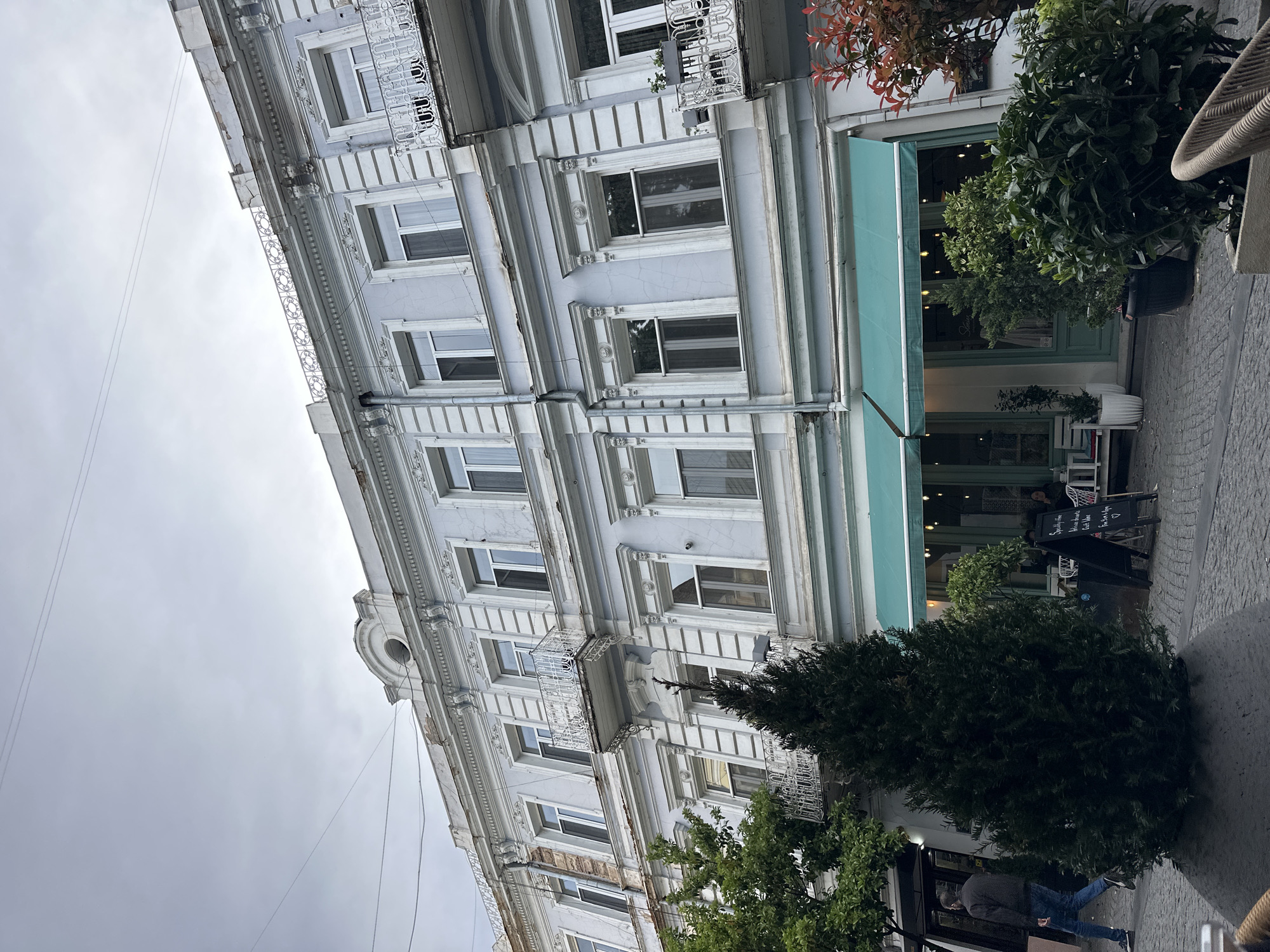 The image depicts a multi-story building in Tbilisi, Georgia. The architecture is ornate, with decorative moldings, window frames, and small balconies with wrought iron railings. The building's facade is painted in a light greyish-blue color. A green awning extends over the entrance of a café or restaurant, which has dark green framed doors. In front of the establishment, several people are present; one man walks by on the sidewalk, and two people are near a blackboard menu display. Various potted plants and greenery are situated near the entrance. The sky is overcast and grey.