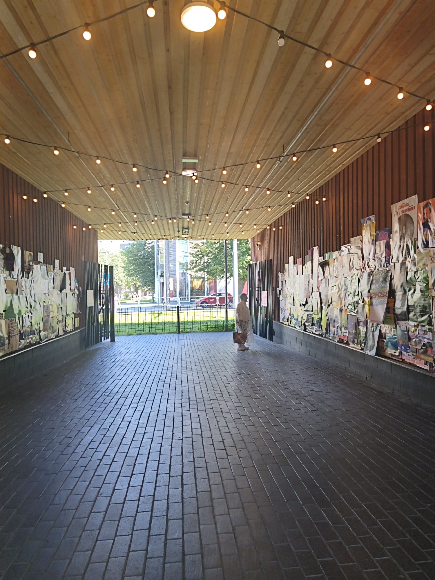 The image captures an outdoor covered walkway with a tiled floor and a wooden ceiling adorned with strings of lights. On both sides of the walkway, walls are covered with numerous posters. A single person, dressed in light-colored clothing and carrying a bag, is visible walking in the middle of the walkway, facing away from the camera. Beyond the walkway, a street with trees, buildings, and a red car can be seen. The setting appears to be a public area in the city of Tallinn, Estonia, likely a passageway leading to or from a building, possibly an exhibition space or venue given the abundance of posters. The lighting suggests it is daytime, though the covered nature of the walkway creates a somewhat dim environment.