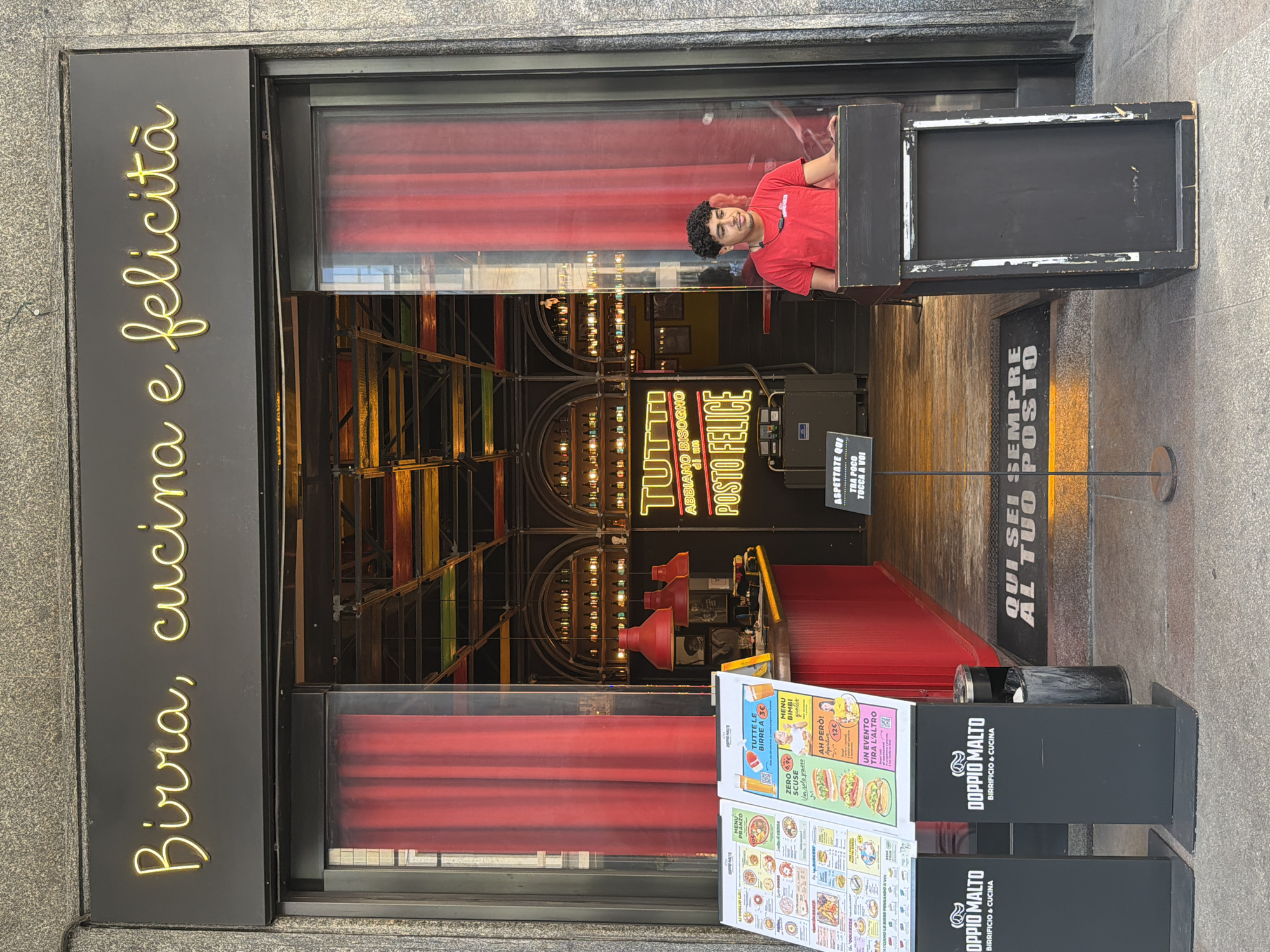 A vertical shot shows the facade of a restaurant called "Doppio Malto," located in Milan, Italy. The restaurant's name is displayed on the sign, along with the phrase "Birra, cucina e felicità" in yellow neon lettering. A man in a red shirt stands inside the restaurant, looking out through the window. The interior of the restaurant is visible through the window, revealing a modern design with various lights and decorative elements. Red curtains are visible behind the man and throughout the restaurant. A menu stand with an array of advertisements is positioned outside. A black trash receptacle is situated nearby. A sign on the floor reads "Qui sei sempre al tuo posto." The scene takes place during daylight hours.
