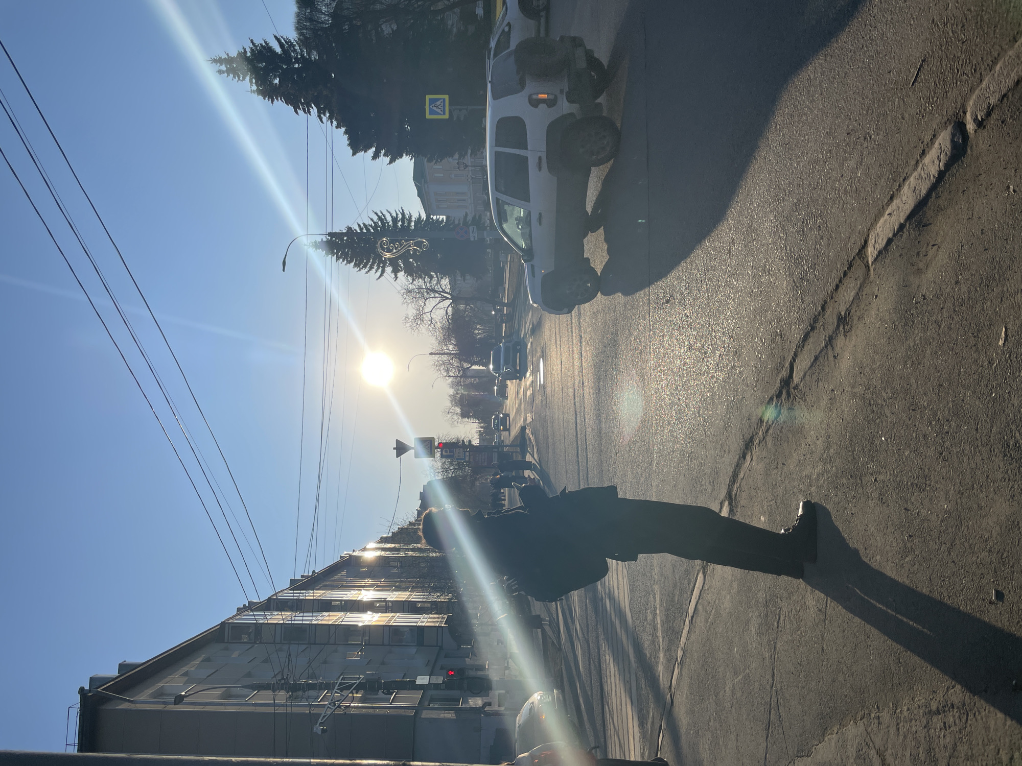 The image, displayed rotated 90 degrees counter-clockwise, depicts a sun-drenched city street in Izhevsk, Russia. If viewed correctly, the scene would show a street running horizontally with buildings on the left and the sun low in the sky on the left side of the frame, suggesting either early morning or late afternoon. The low sun angle creates long shadows, and the bright light causes significant lens flare and reflections off buildings and the wet-looking asphalt road. The sky is clear and blue.
In the mid-ground on the right side of the street, a person is silhouetted against the bright light, appearing to be walking or standing. Their long shadow stretches across the pavement to the right. The asphalt road shows cracks and glistens from the sun's reflection. Several vehicles are visible, including a prominent white SUV or truck in the mid-ground, and other cars further down the road.
On the left side of the street, multi-story buildings with numerous windows reflect the intense sunlight. Overhead power lines crisscross the sky. Trees, some appearing bare of leaves and others evergreen, line parts of the street.
Visible street furniture includes a street lamp. Traffic lights, one of which appears red, are also present. Road signs include a yellow pedestrian crossing sign depicting a black figure of a person walking, and a red 'P' parking sign further in the distance. The scene captures typical urban activity under clear, sunny weather.