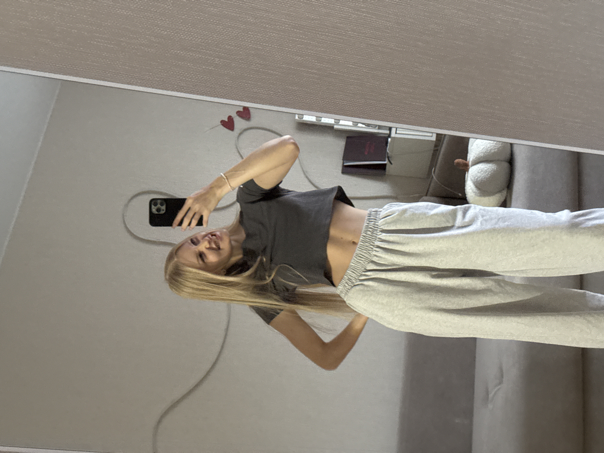 A young Caucasian woman with long blonde hair is taking a mirror selfie in a room. She is wearing a dark grey cropped t-shirt and light grey sweatpants. She is smiling and has her left hand on her hip while holding a smartphone in her right hand. The background shows a light grey wall with a white decorative swirl, two red heart decorations, and a shelf with books and other items. A couch and a plush pumpkin pillow are visible in the reflection on the left side of the mirror. The overall setting suggests a casual indoor environment.