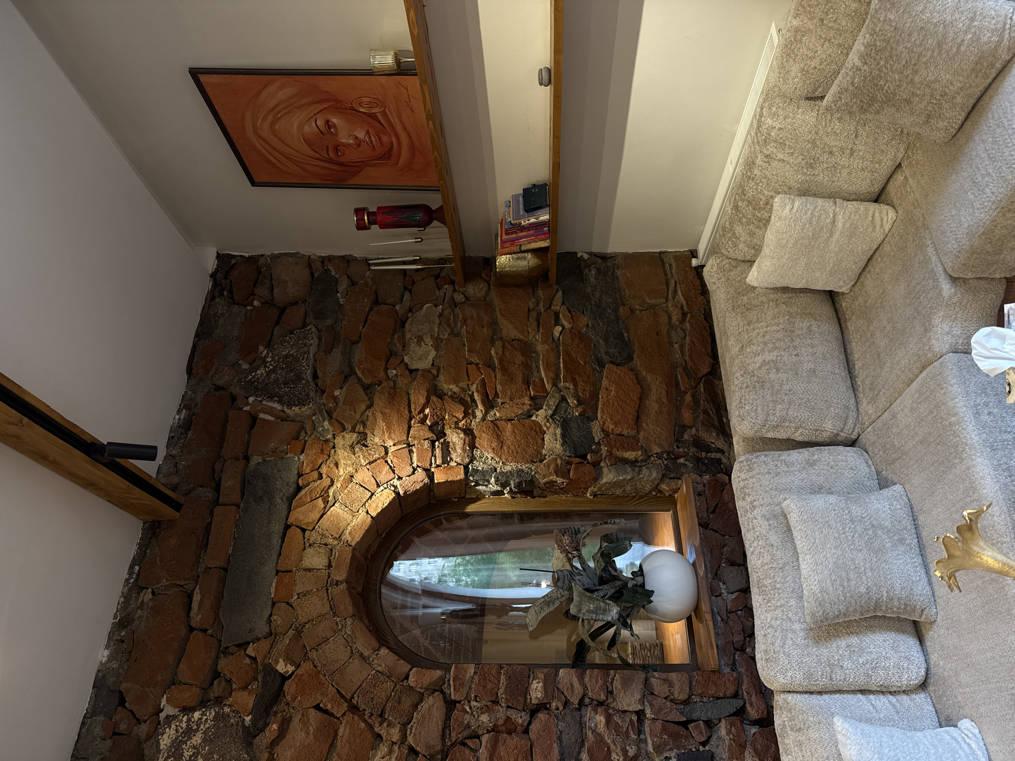The image depicts an interior space with distinctive features, likely a living room or similar area. The dominant element is a stone wall constructed from irregularly shaped, reddish-brown rocks. Integrated into this wall is an oval window with a wooden frame, showcasing a view of greenery and sky, suggesting an exterior landscape. In front of the window sits a decorative plant with large leaves and a spherical, white vase or lamp.
Hanging on the wall, above the stone structure, is a framed painting of a person with a head covering. A shelf is mounted on the wall near the painting, containing books and a decorative red and green object.
To the right of the stone wall, a large, light gray sectional sofa is positioned. It includes several cushions and appears to be made of a textured fabric. A golden decorative object is partially visible on the sofa.
The setting has some similarities to architectural styles often found in Yerevan, Armenia, which are known for the use of local stone in construction and design elements, such as the rock wall, and unique designs. However, without additional information, the precise location cannot be verified.