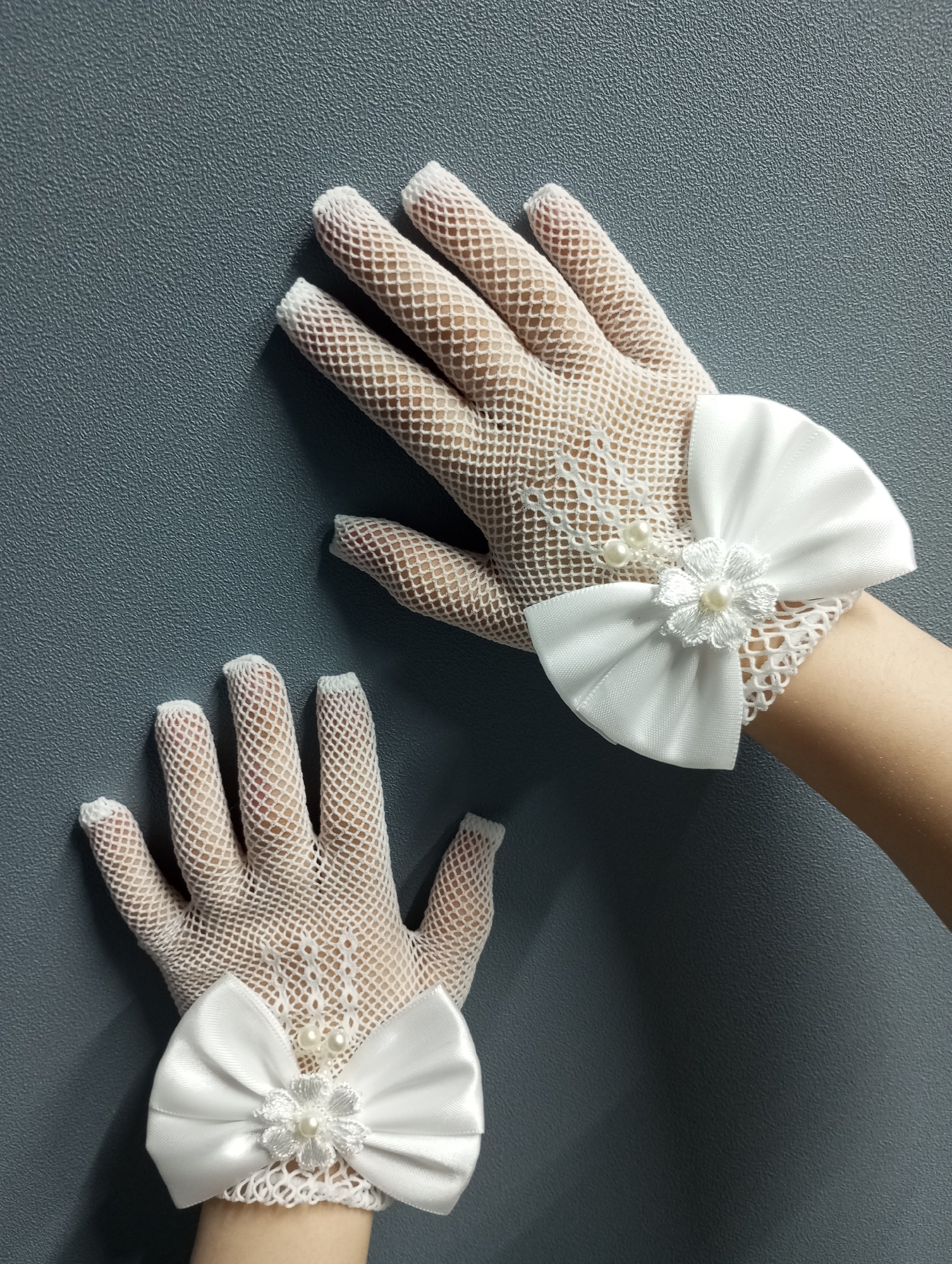 The image displays two hands wearing white fishnet gloves adorned with white satin bows. Each bow features a pearl-centered flower and three pearl embellishments. The hands are positioned against a matte grey background. The setting is not discernible and is not definitively located in Pervomaysky, Russia.