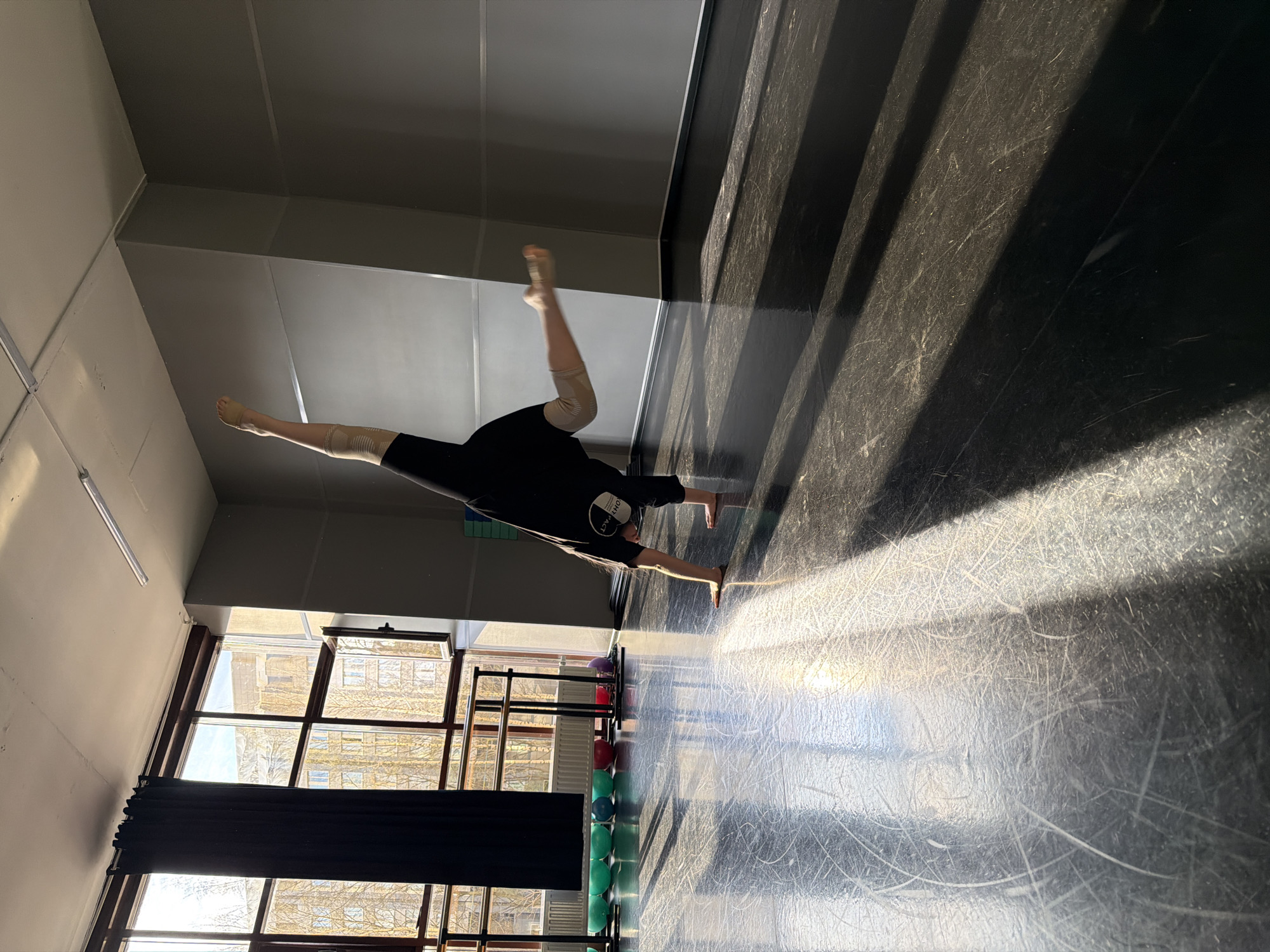 A dancer in a black shirt and beige leggings executes a handstand in a dance studio, one leg extended forward and the other bent upward. The studio has a dark, striated floor and light gray walls. Light streams in from windows behind a black curtain, illuminating the floor. A barre with colorful balls rests along the wall. The location is Saint Petersburg, Russia.