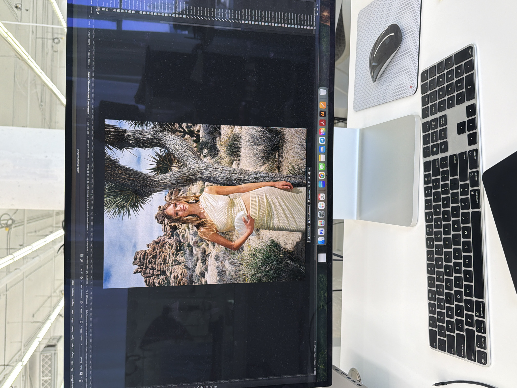 A medium shot of a monitor with a digital image displayed, positioned on a white desk alongside computing peripherals. The monitor screen displays an Adobe Photoshop workspace with multiple files open, including images. An image of a blonde-haired woman wearing a dress is visible within one of the Photoshop files. The backdrop of the image appears to be a desert landscape with rock formations and Joshua trees.
A wireless Apple Magic Mouse and an Apple Magic Keyboard with a numeric keypad are situated to the right of the monitor. The environment around the desk is blurry, with white industrial elements visible, suggesting an indoor setting. The image is taken indoors in a building of Los Angeles.