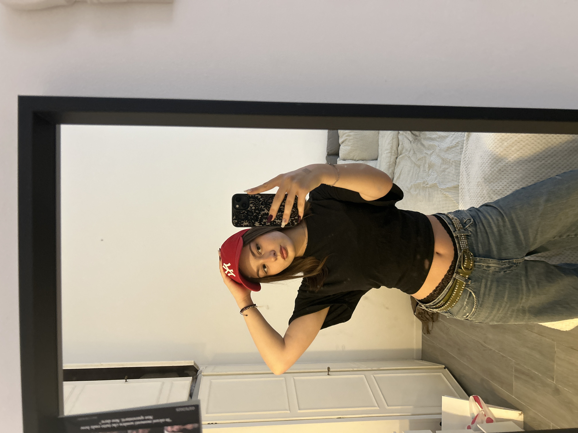 A young woman is taking a selfie in a mirror. She is wearing a black crop top, loose blue jeans, a red baseball cap, and multiple belts. She is holding a smartphone with a black patterned case. The reflection in the mirror also shows part of a bed with white and grey bedding and a white cabinet below. It is not possible to determine if the location is Milan, Italy based on the image alone.