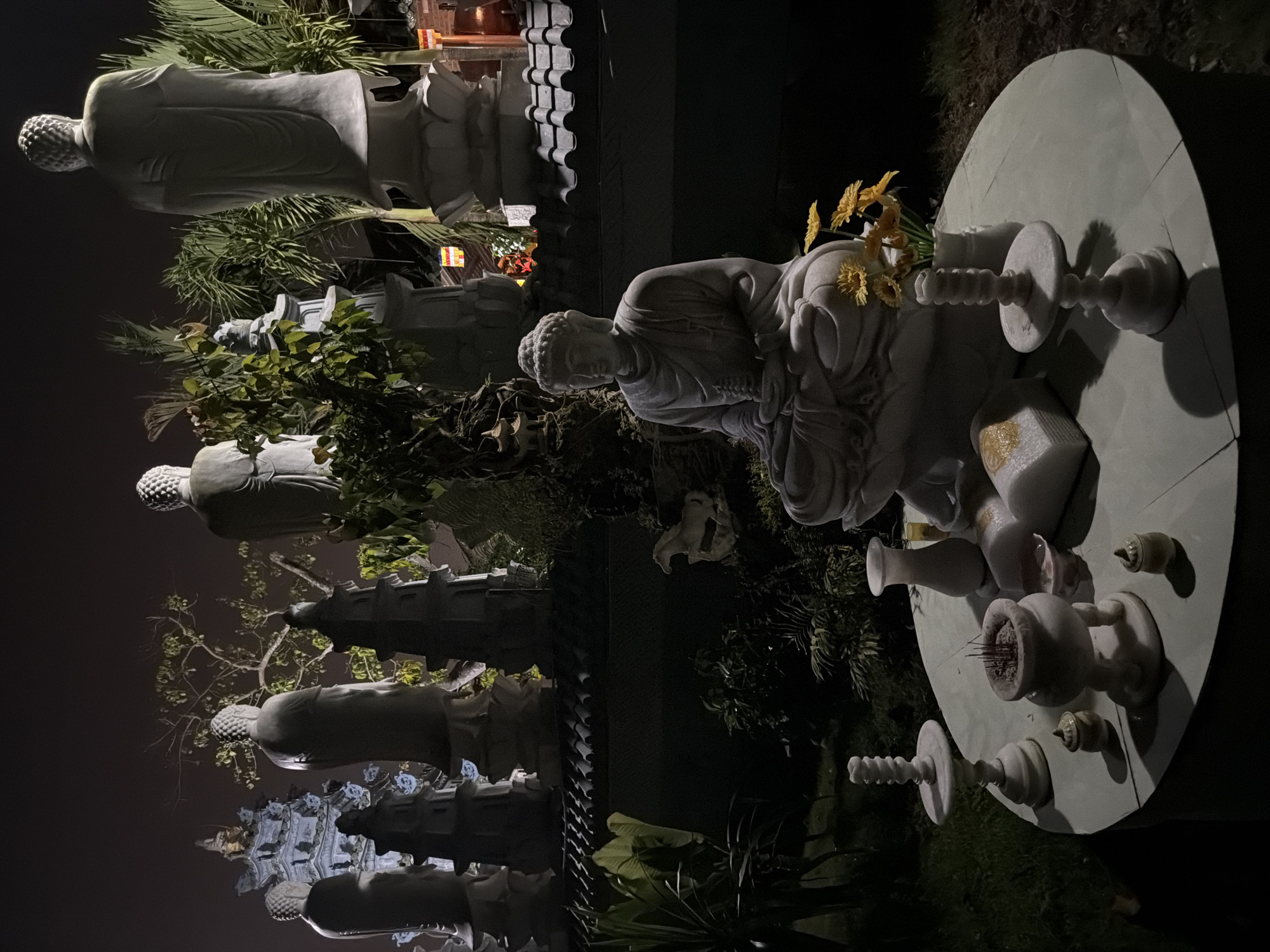 The image shows a nighttime view of a Buddhist temple complex, likely in Đà Nẵng, Vietnam. In the foreground, a round stone table supports various ritual objects, including a vase of yellow flowers, incense holders with incense sticks, and other decorative elements. A kneeling Buddha statue with intricate details is positioned near the table. In the background, a row of standing Buddha statues are visible, lined up in the temple grounds. The statues and temple structures are illuminated, contrasting with the dark night sky. Lush greenery and trees surround the area, enhancing the spiritual ambiance.