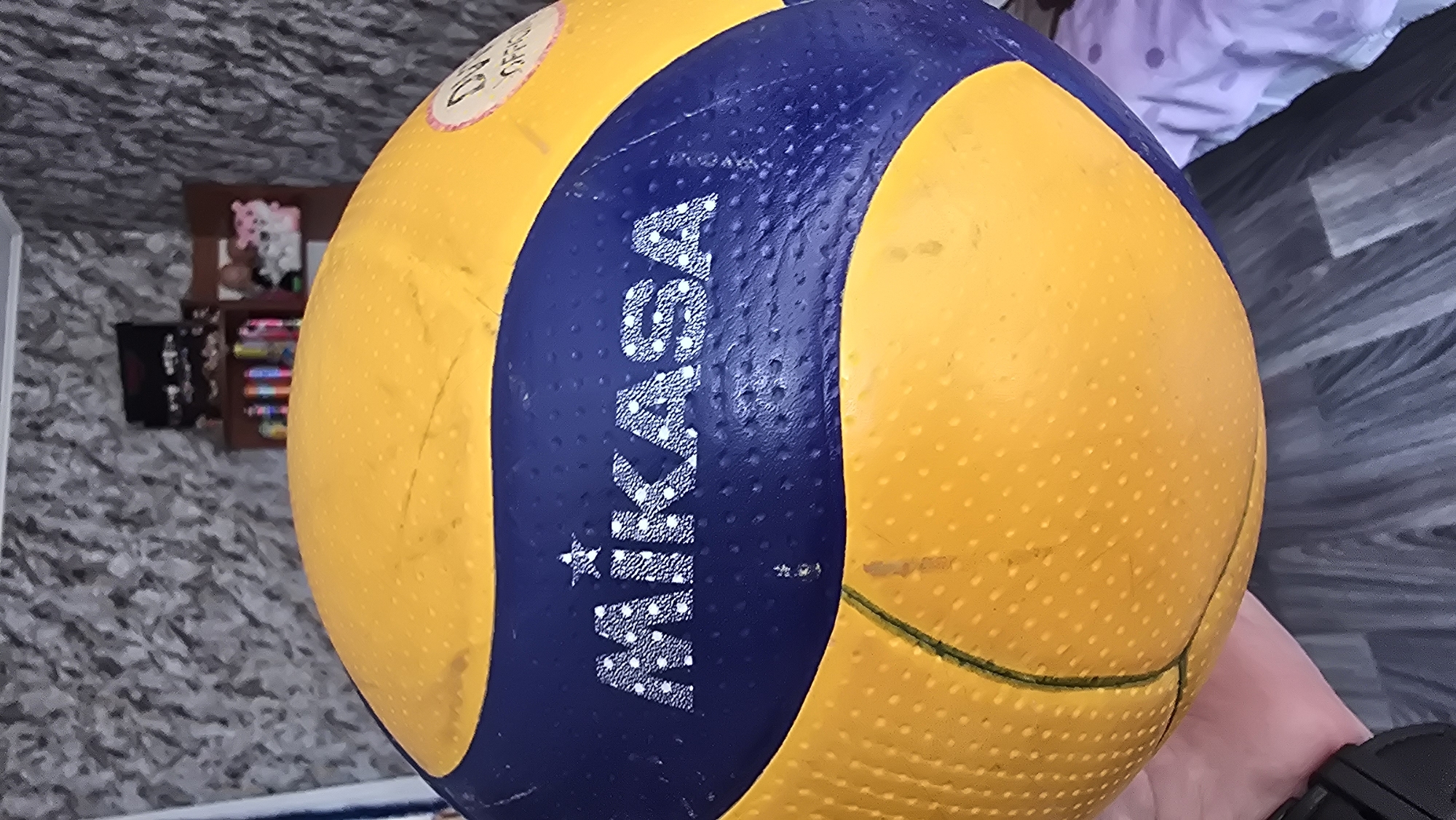 A yellow and blue Mikasa volleyball is held in a person's hand, positioned in front of a wall with patterned wallpaper. In the background, a shelf unit contains various items, including what appear to be toys and decorative objects. The floor is covered with dark, wood-patterned laminate flooring. The scene is indoors, likely a room in a residential setting within Novohuivynske, Ukraine.