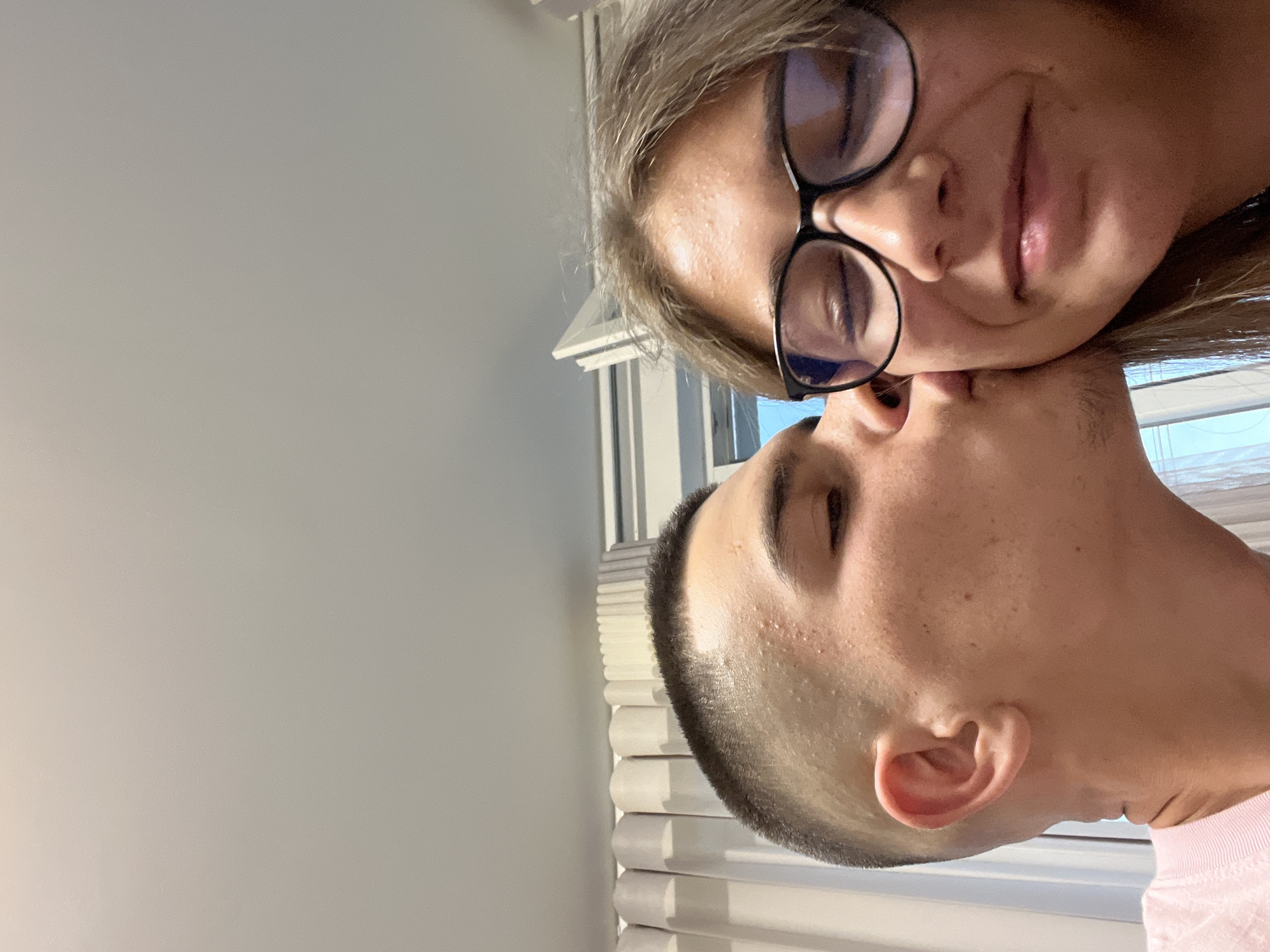 A close-up, portrait-oriented indoor photograph captures two individuals in an intimate moment within the city of Karavelovo, Bulgaria. On the left side of the frame, a male with a very short, faded haircut and light complexion is depicted with his eyes closed, in the act of kissing the right cheek of a female. He wears a light-colored, ribbed t-shirt, the collar of which is partially visible. The female, positioned on the right side of the frame, has light brown hair and wears black-framed eyeglasses; her eyes are closed, and she displays a subtle smile on her face, receiving the kiss. Both individuals exhibit minor skin blemishes on their foreheads and cheeks. The background features light-colored, pleated curtains on the left and center, partially obscuring a window with a white frame structure on the right. The upper portion of the image reveals a plain, light-toned ceiling or wall.