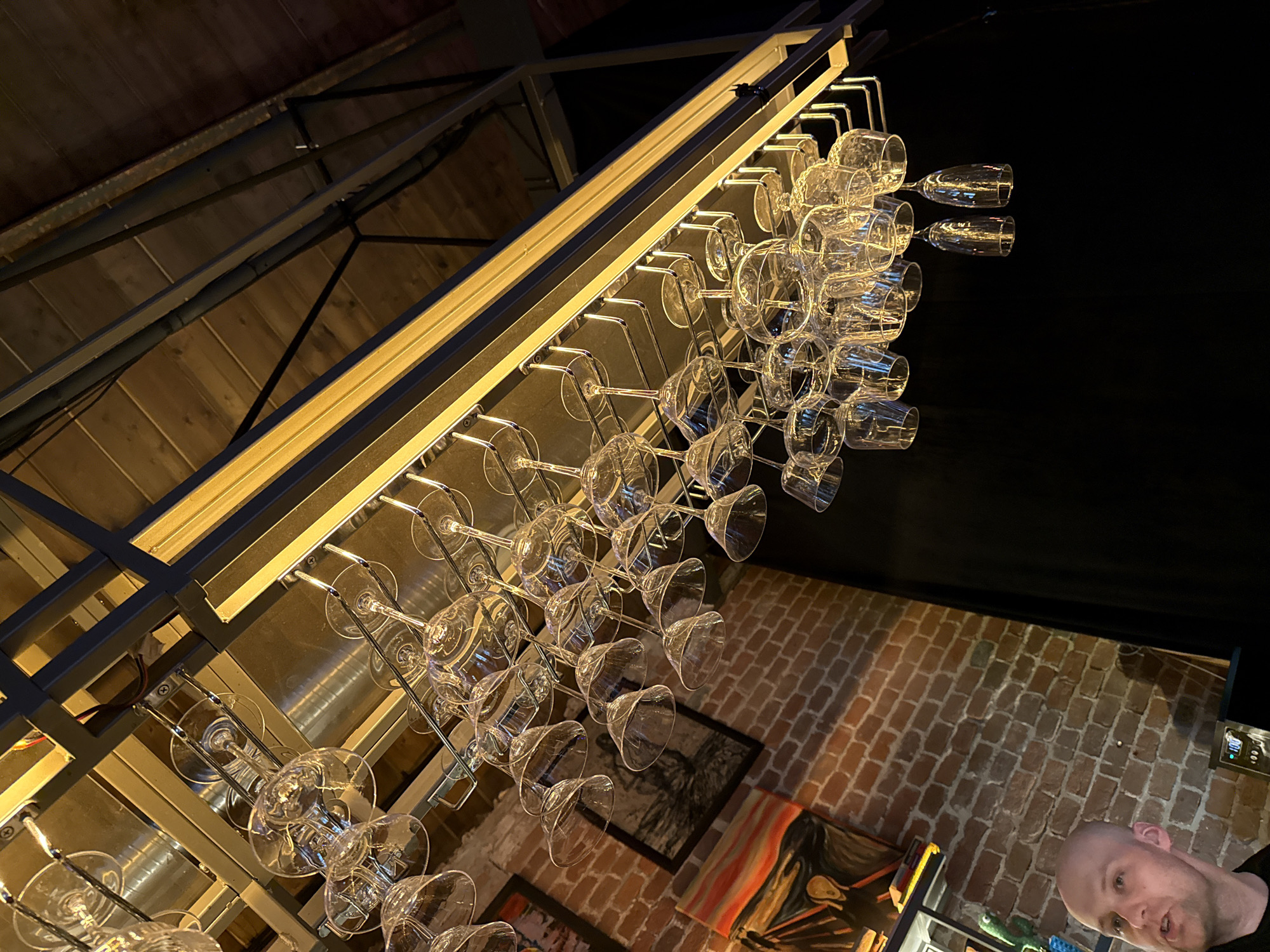 The image depicts a bar or restaurant interior. A metal rack holding rows of wine glasses hangs overhead. The ceiling appears to be constructed of wood planks and metal supports. One brick wall is visible, and a person's head is partially visible at the bottom right corner of the image. Art hangs on the brick wall. There is no indication from the image that the location is in Moscow, Russia.