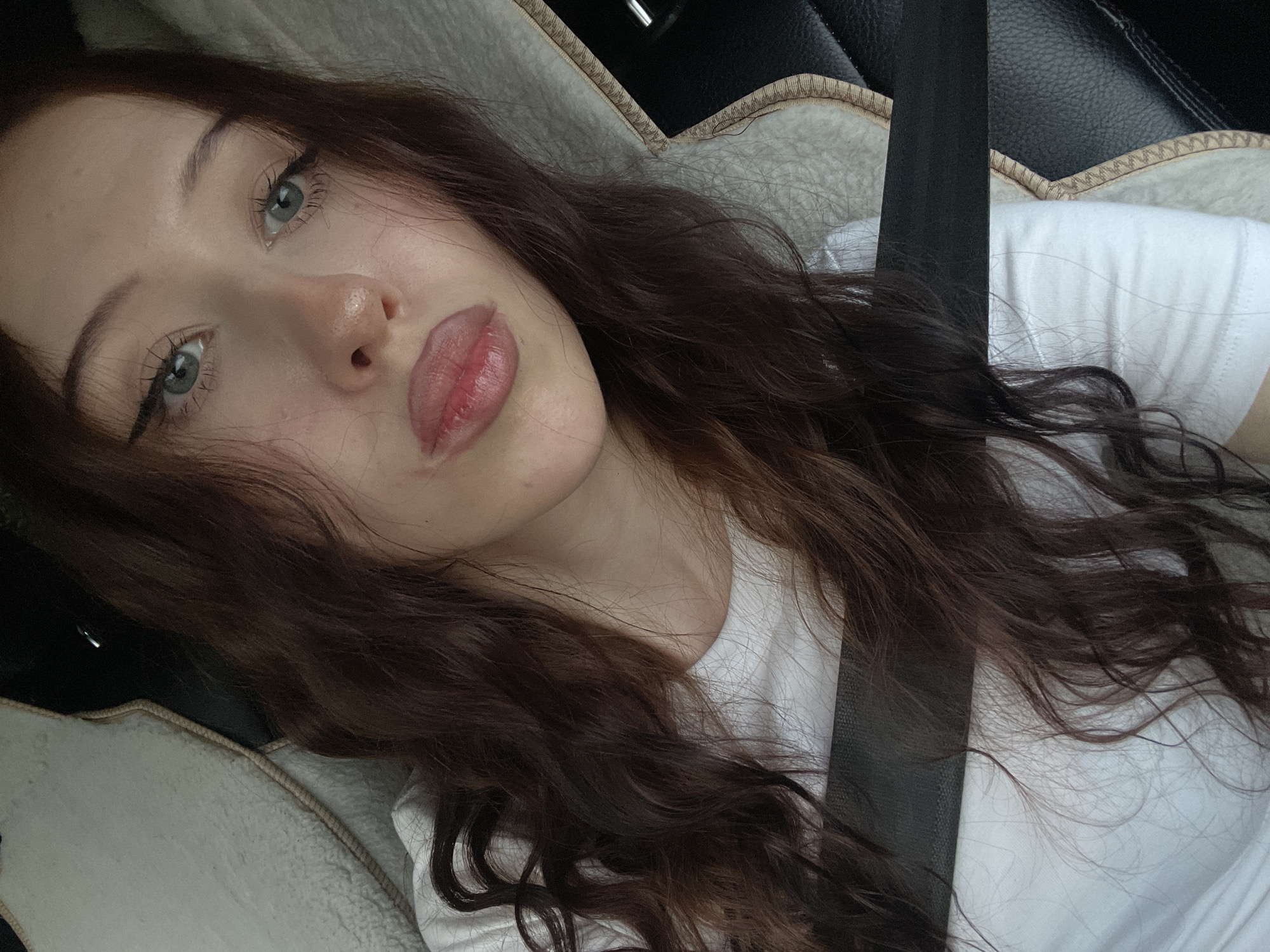 This media file is a close-up selfie of a young woman taken in a car. The woman has long, dark brown, wavy hair and light blue eyes. She is wearing a white t-shirt and appears to be wearing light makeup, with eyeliner and lip gloss visible. The seatbelt is fastened across her chest. The background shows a car interior, with textured upholstery and a black leather seat. The lighting suggests it might be daytime, possibly late afternoon, based on the soft, natural light. The woman's expression is relaxed and she appears to be posing for the photo, looking directly at the camera. There is no visible text in the image. The location context is Votkinsk, Russia.