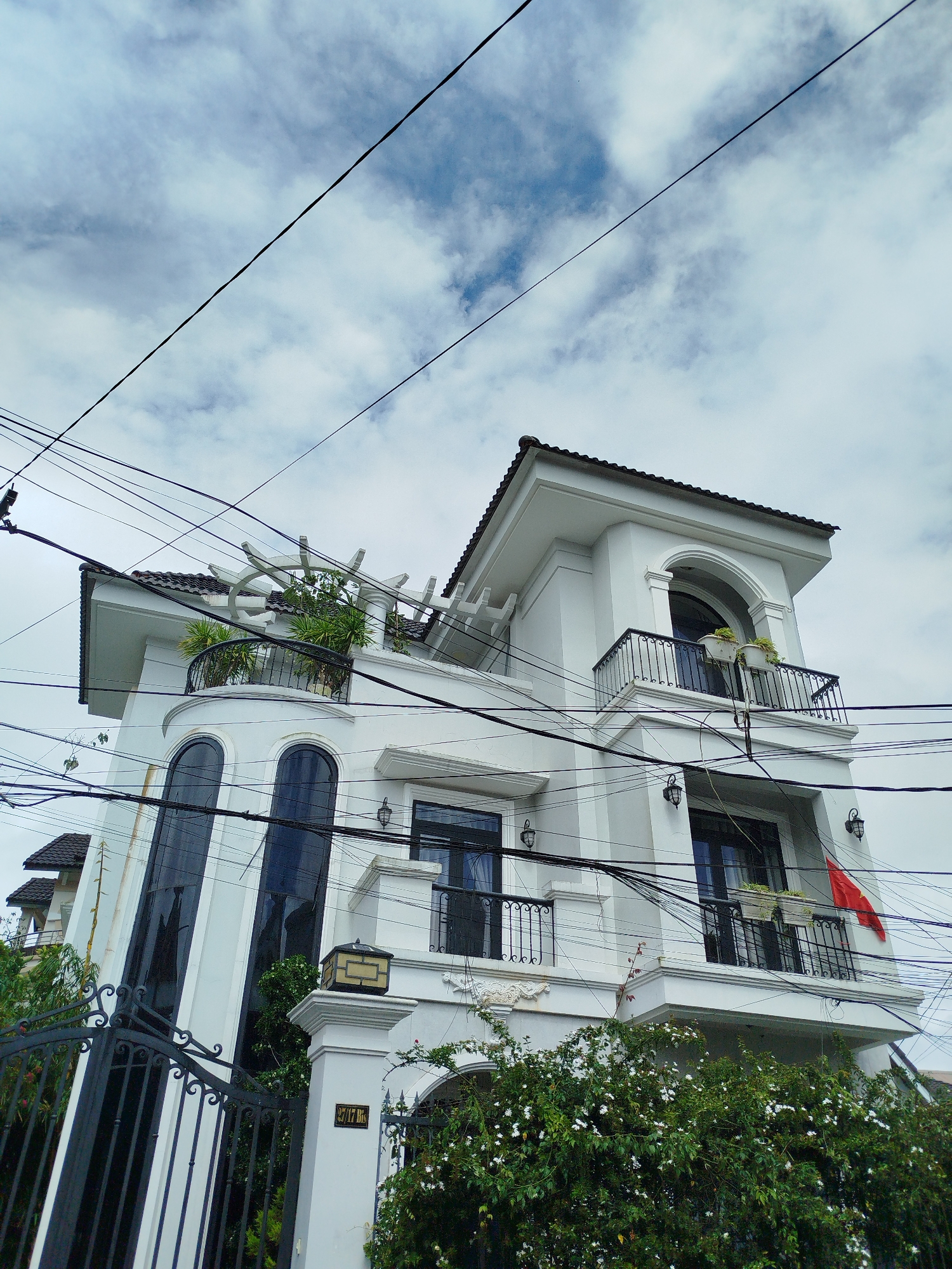 This is a low-angle, full shot of a modern white multi-story house in Da Lat, Vietnam, situated under a cloudy sky crisscrossed with numerous overhead electrical wires. The house has several balconies with black metal railings and potted plants. One balcony features an arched window above it, and another has a red flag hanging from it. The architecture is characterized by clean lines, large dark-paned windows, and a tiled roof. The front of the property includes a black wrought-iron gate and a wall with a plaque bearing the numbers "27/17 BK". Dense green foliage with white flowers is visible in the foreground, partially obscuring the lower section of the house and gate. The overall impression is of a residential building in an urban or suburban setting with abundant greenery and visible utility infrastructure.