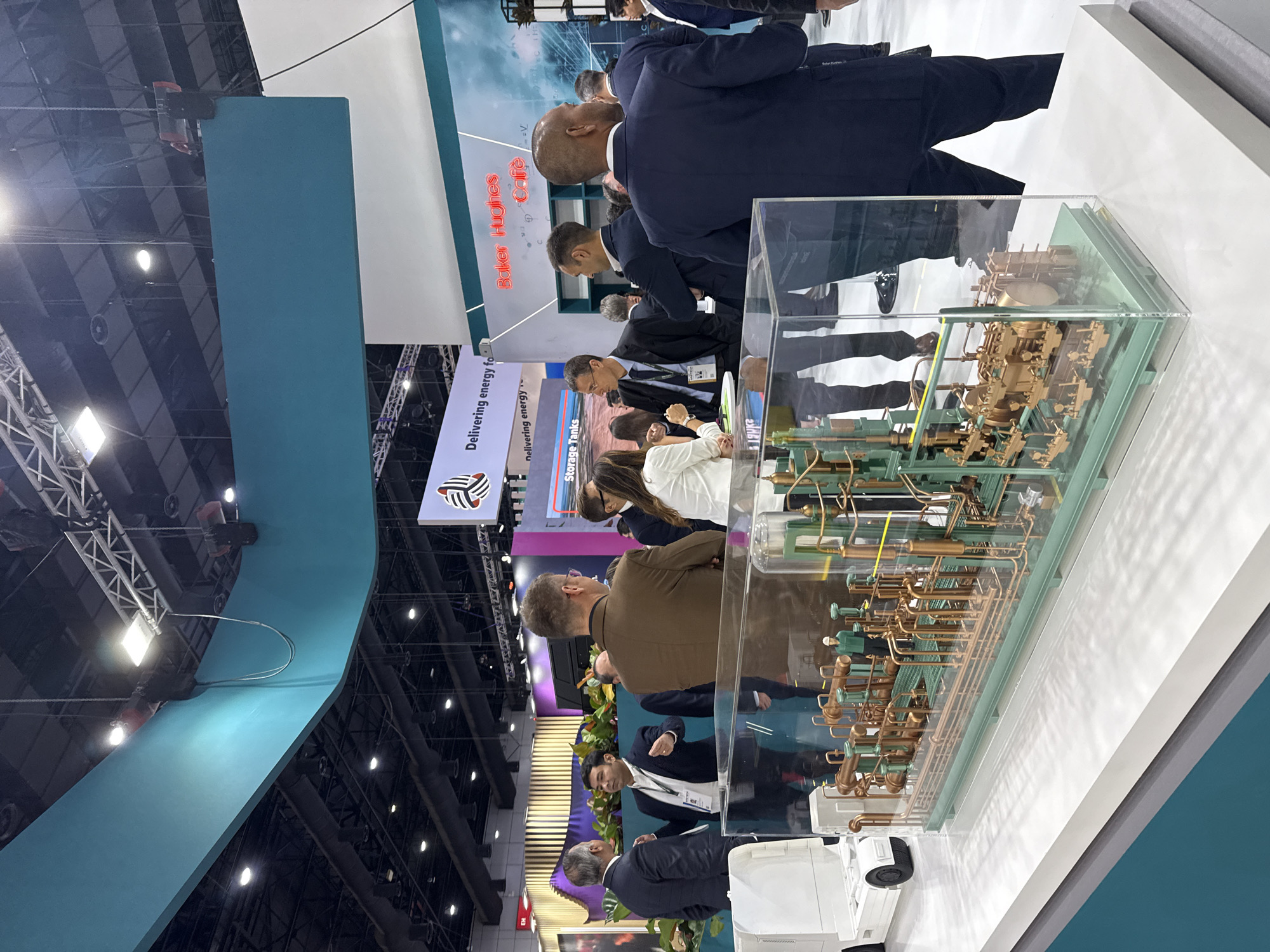 The image shows an indoor exhibition hall, likely part of a trade show or convention center. A group of people, mostly men dressed in business attire such as suits and blazers, are gathered around a display. The display features a detailed model of a complex industrial processing unit with numerous pipes and machinery, encased in clear acrylic. A white miniature truck is also part of the display.
In the background, there are exhibit booths with company logos and signage. One prominent sign reads "Baker Hughes Cafe" in neon red letters. Another sign visible further back has a logo and text that seems to say "Delivering energy for..." and "Storage Tanks". The ceiling features industrial lighting and structural elements. The overall environment suggests a professional event focused on the energy or industrial sector.
While the image depicts an indoor event, there is no visual information within the image itself that can definitively place the location in Bangkok, Thailand. The scene is generic to exhibition halls worldwide.