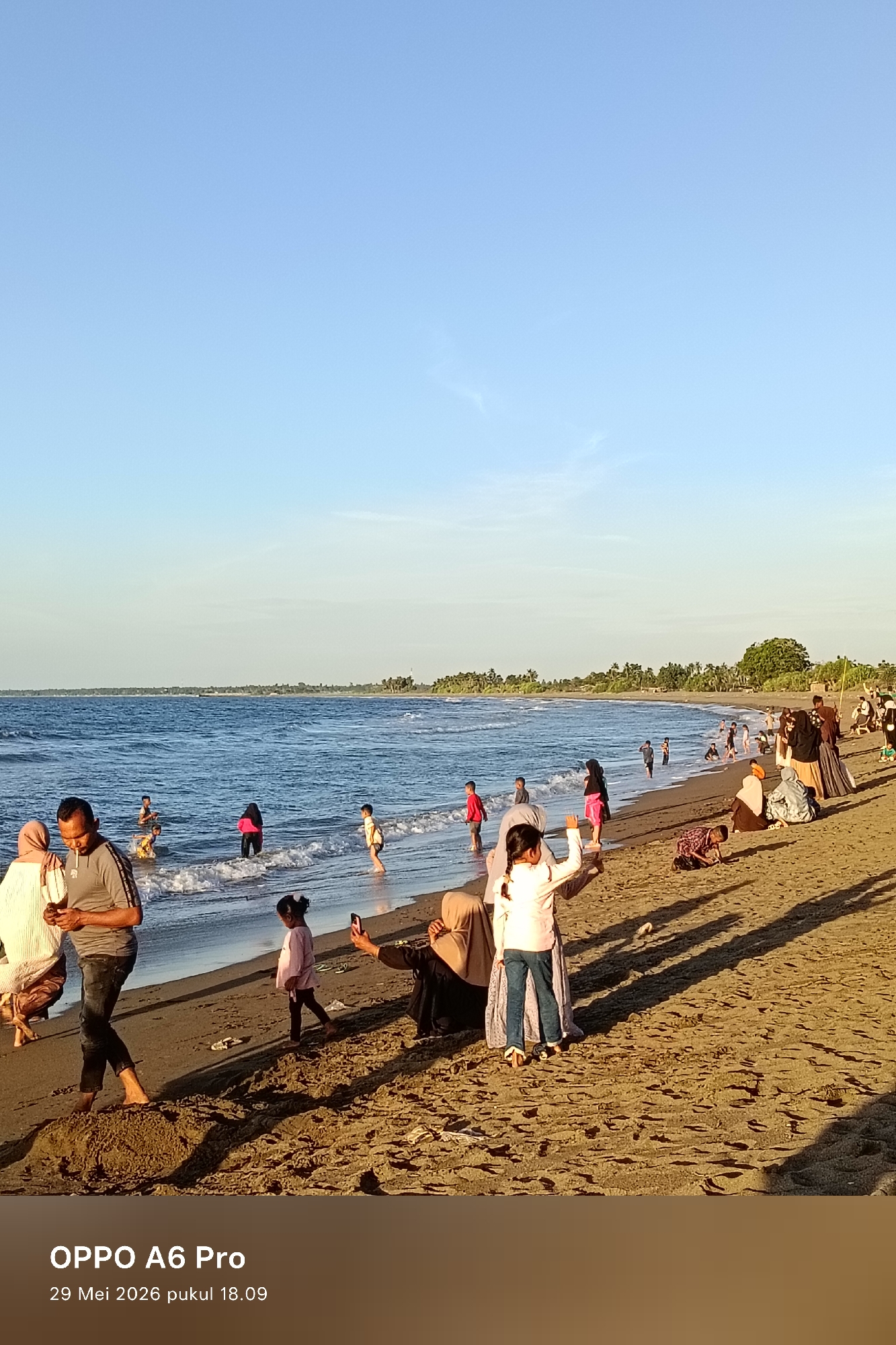 The image shows a beach scene in Rheumbaroh, Indonesia. The sky is clear with a blue gradient. A body of water, likely the ocean, is visible with small waves. Several people are present on the sandy beach. Some are wading in the water, while others are standing or sitting on the sand. A group of trees is visible in the background along the coastline. The lighting suggests it is late afternoon. An OPPO A6 Pro watermark and timestamp "29 Mei 2026 pukul 18.09" are present at the bottom of the image.