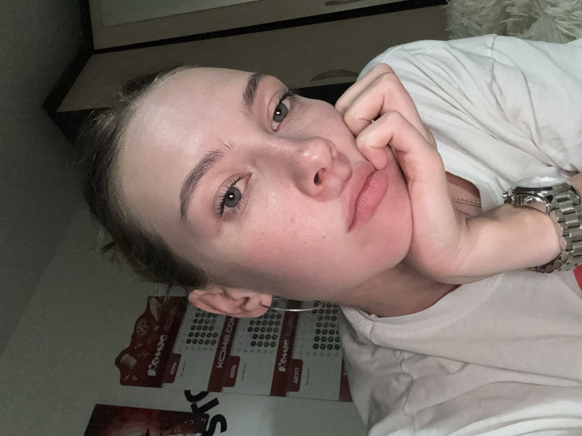A young woman is captured in a close-up, indoor selfie. She has fair skin, light brown hair pulled back, and is wearing a white t-shirt. Her right hand rests on her chin, and she looks directly at the camera with a pensive expression. In the background, several calendars with Russian text and circular date markers are visible, suggesting a domestic or office setting. The overall lighting is dim, contributing to a casual and intimate mood. The presence of calendars with Russian text could indicate the location is in Russia, but the specific city of Perm cannot be confirmed from the visual information alone.