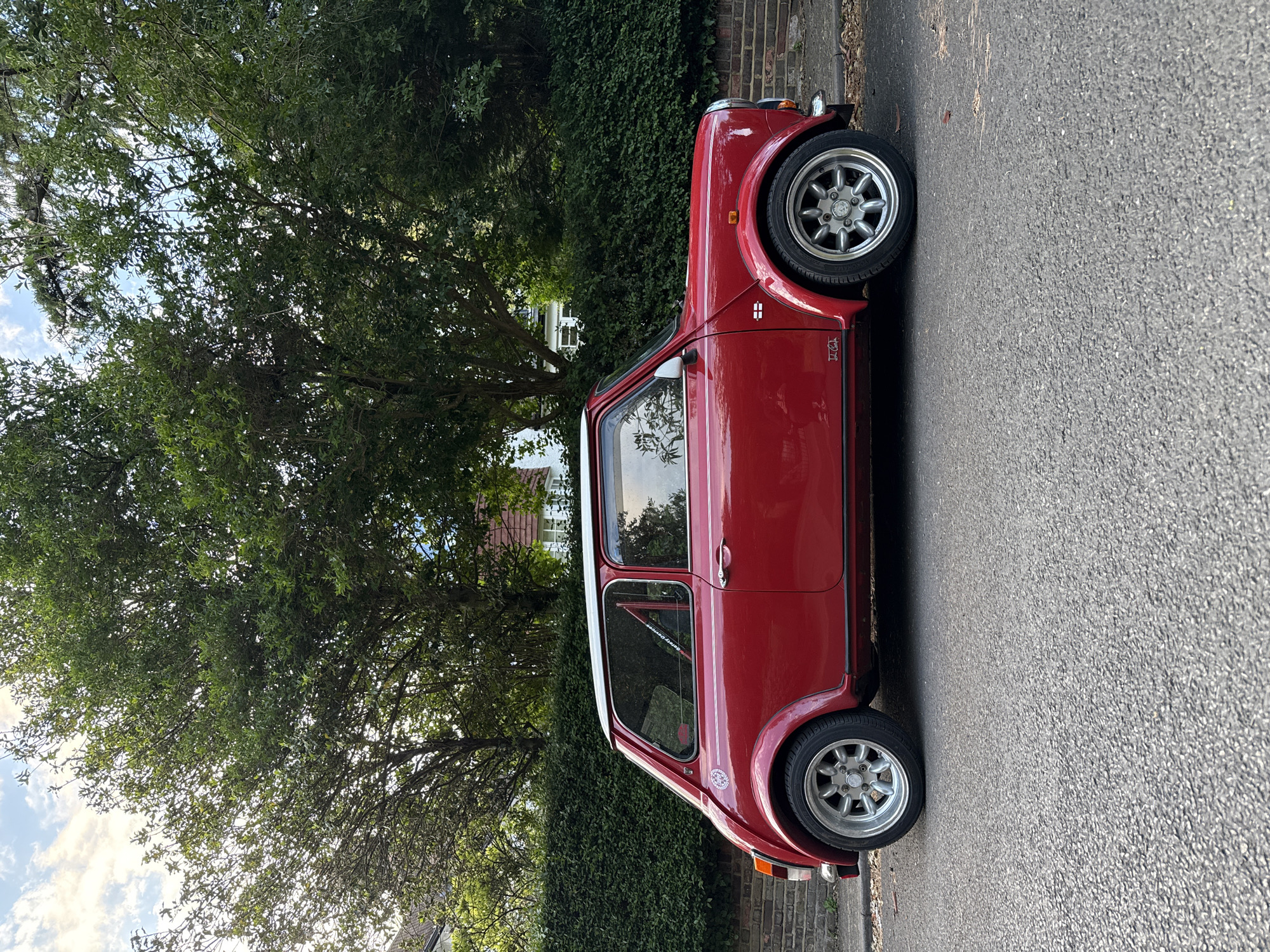 A red classic Mini Cooper car is parked on the side of a road in a residential area. The car has silver wheels and a white roof. There's greenery, possibly a hedge and a tree, in the background. The road appears to be asphalt. The sky is visible through the tree branches. The car model is Austin Mini. The exact location cannot be determined with certainty from the image alone. However, given the architectural style of the buildings and the type of vegetation, it's plausible that the image was taken somewhere in the UK.