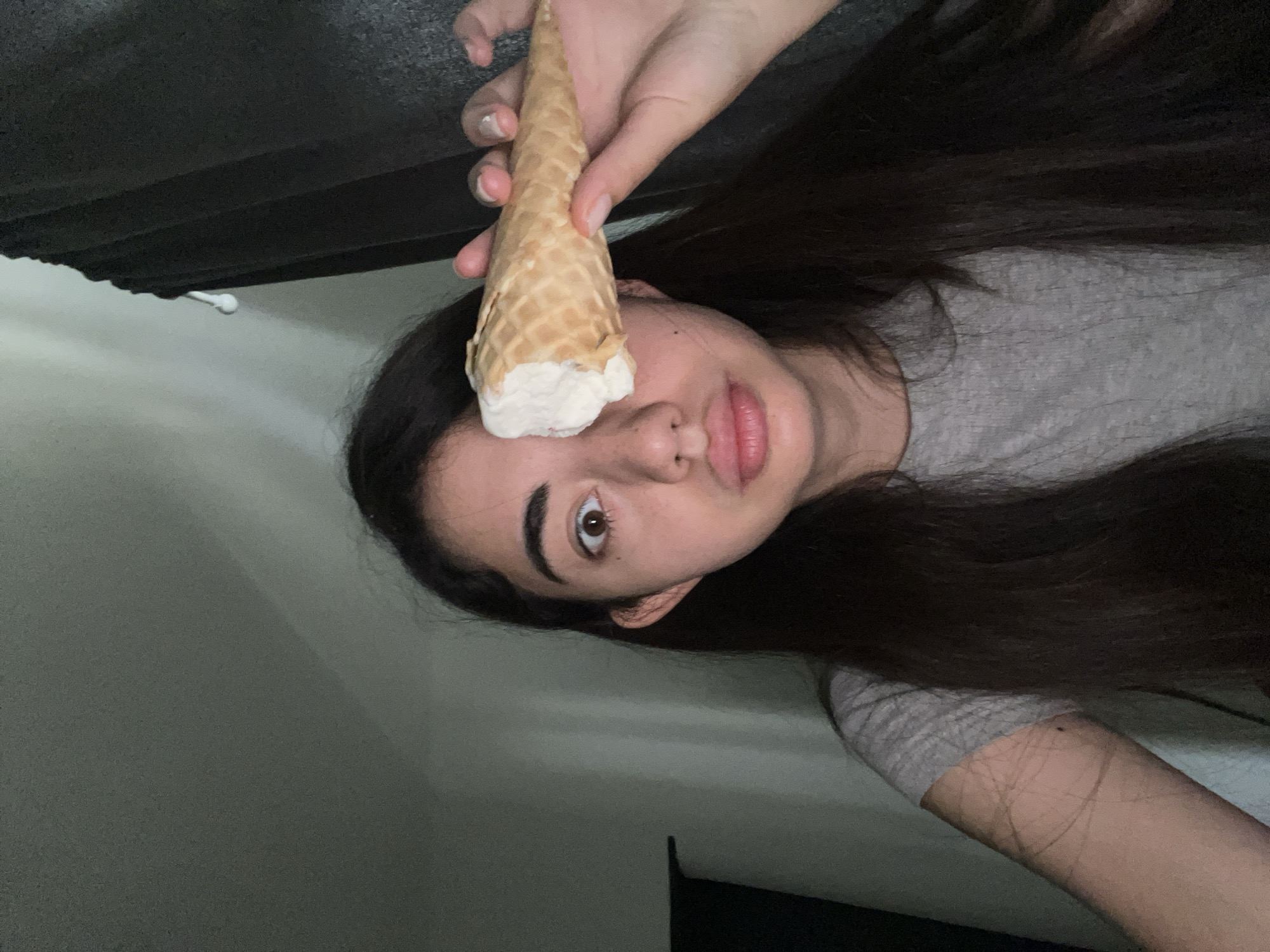 A young person, appearing to be of East Asian descent, is holding a partially eaten ice cream cone in front of their right eye. The person has long, dark hair, brown eyes, and is wearing a grey t-shirt. The background appears to be an indoor setting with a wall and dark curtains. There is no indication of the location in Chotěšov, Czechia. The lighting is dim.